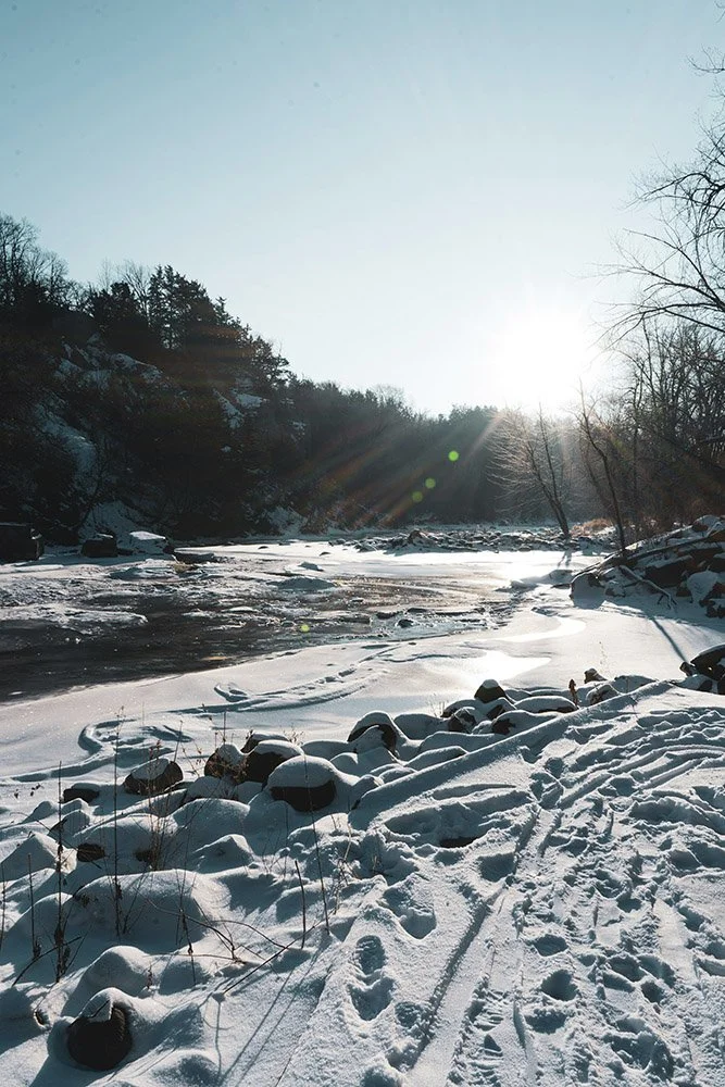 Snow-covered riverbank with footprints, rocks, and leafless trees under a bright sun.