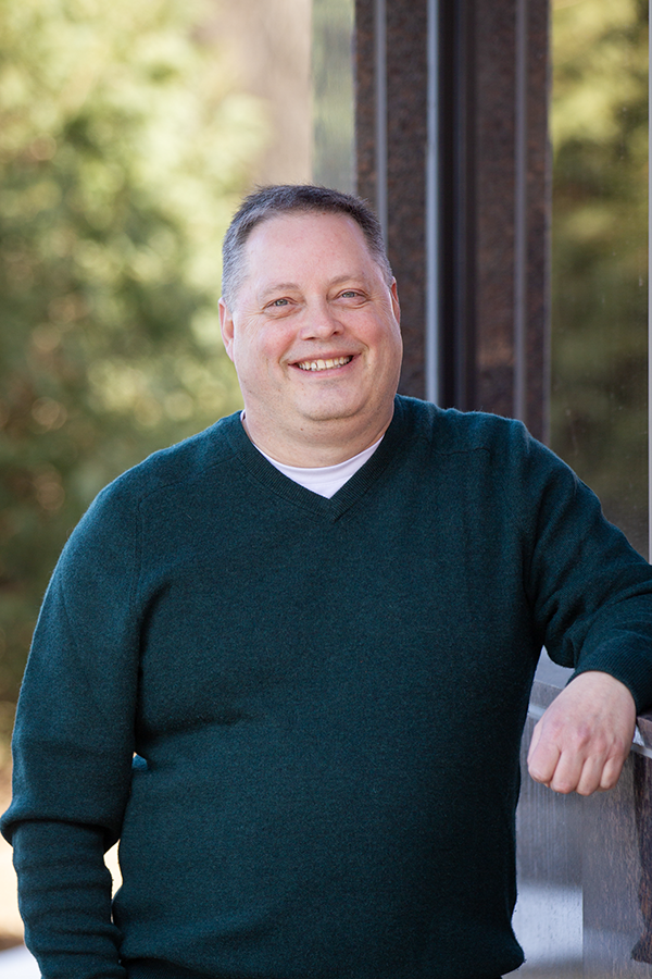 A smiling man with short dark hair, wearing a dark teal sweater and a white shirt, standing outdoors next to a large window with trees in the background.