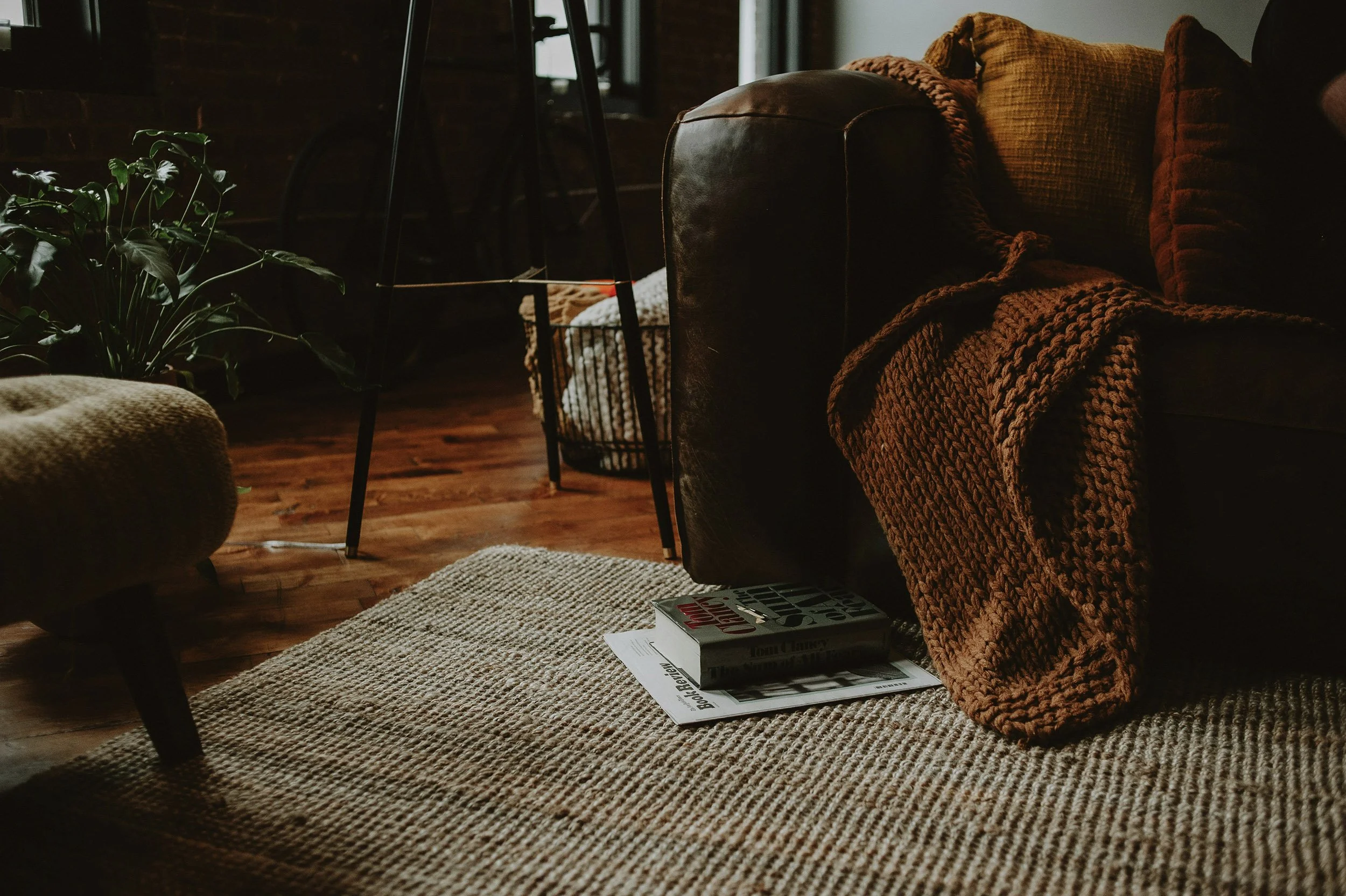 Cozy living room corner with a dark leather sofa, a chunky knit blanket, a potted plant, a woven basket, and a carpeted floor, with books on the floor.
