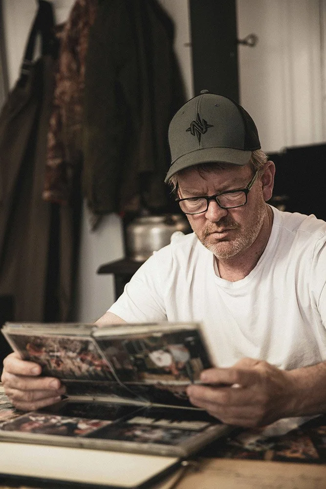 A man wearing glasses and a gray baseball cap is sitting at a table, reading a large photo album.