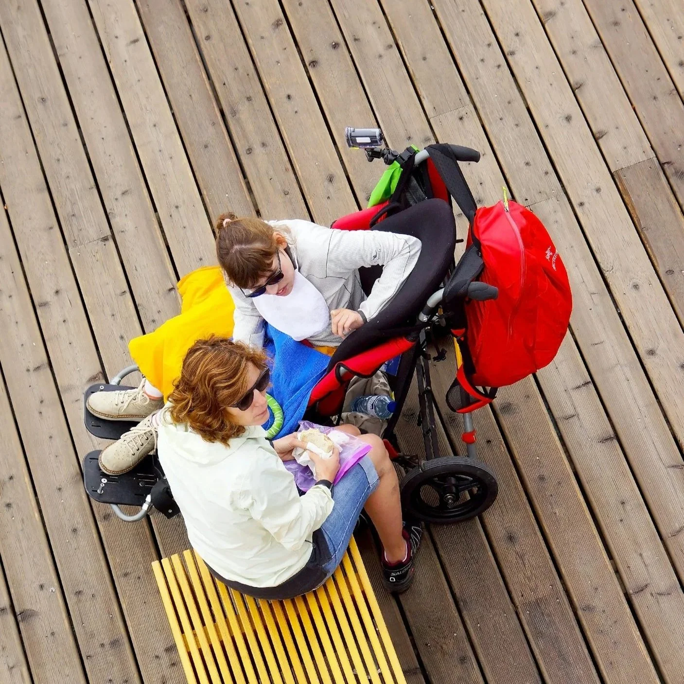 Two women with sunglasses sitting on a bench and a person in a wheelchair on a wooden deck. The woman in front is eating a sandwich and the woman behind is sitting with a colorful blanket over her lap.