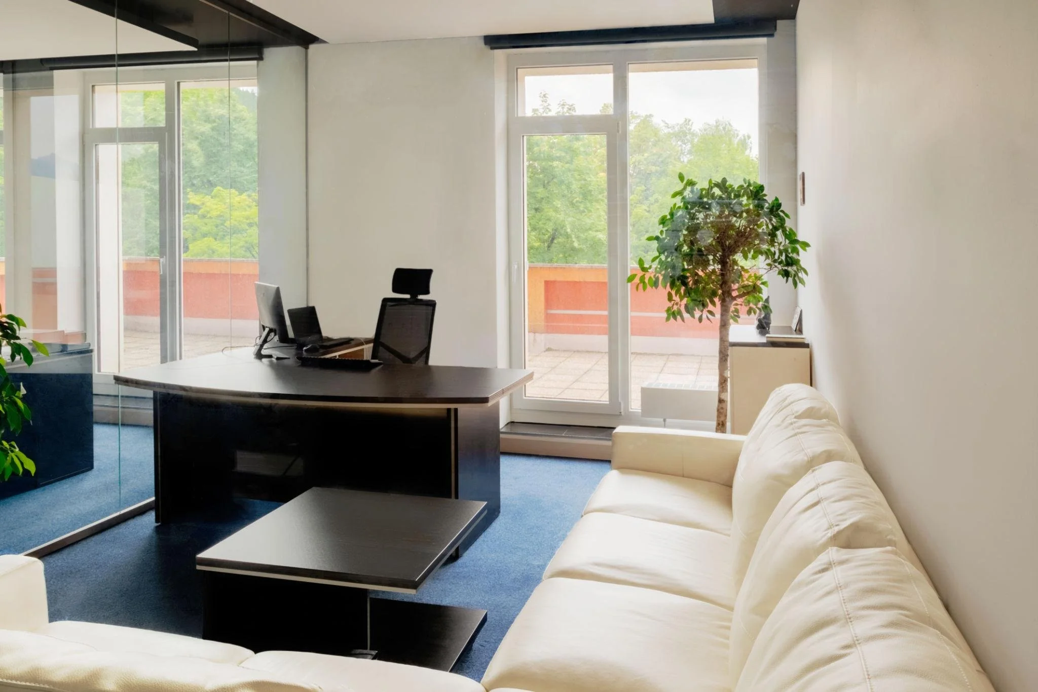 An office with a white sofa, a desk with two computers, a chair, and a large window/door with a view of a balcony and green trees outside.