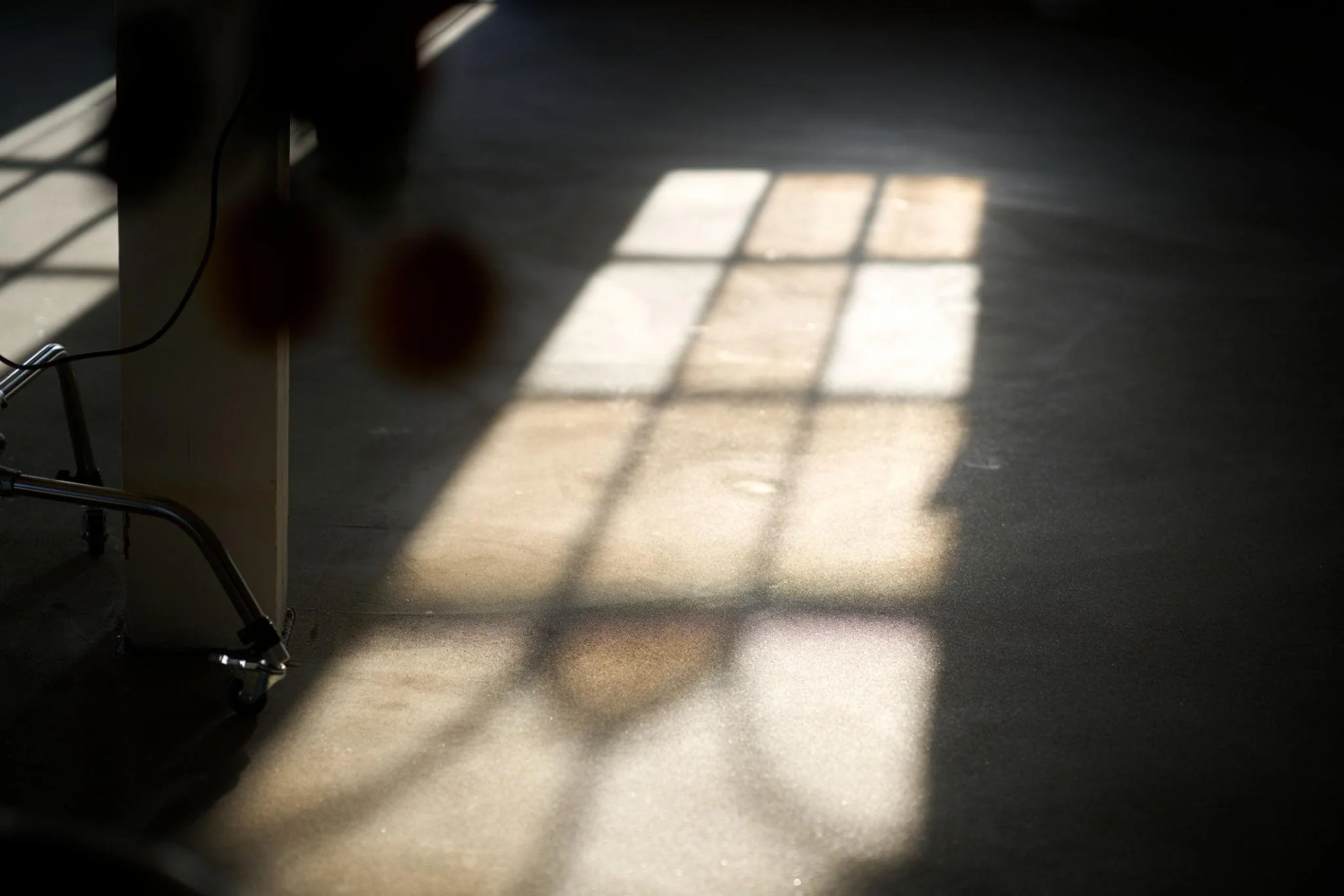 Sunlight coming through a window creates a grid shadow on a dark floor in a room, with part of a metal chair visible on the left side.
