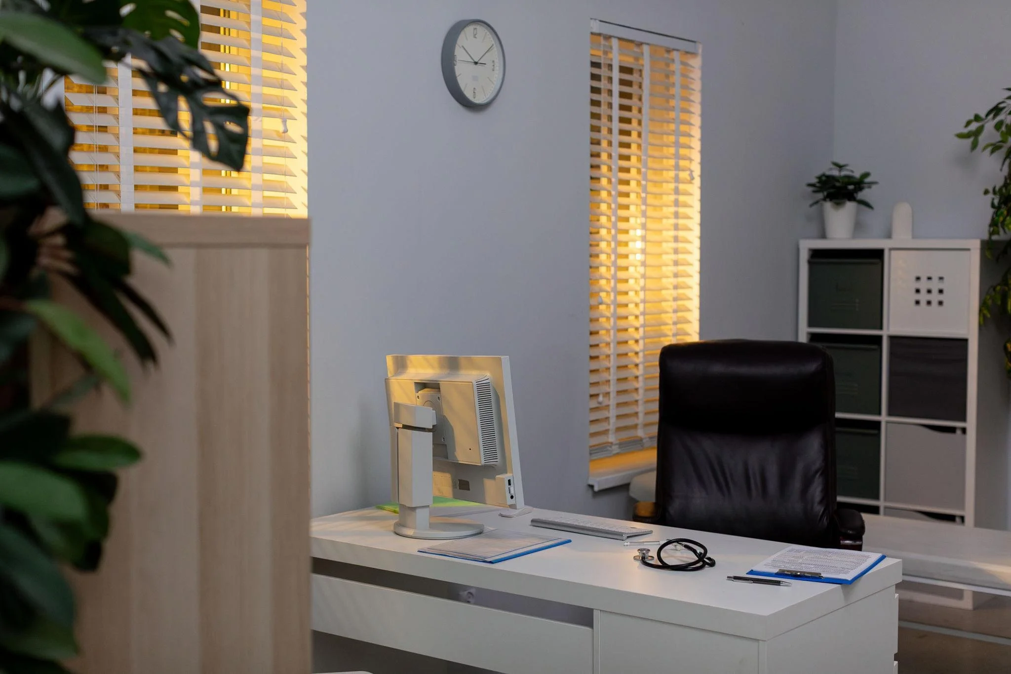 Office workspace with a white desk, computer monitor, clipboard, stethoscope, and office chair, with window blinds and wall clock in the background.