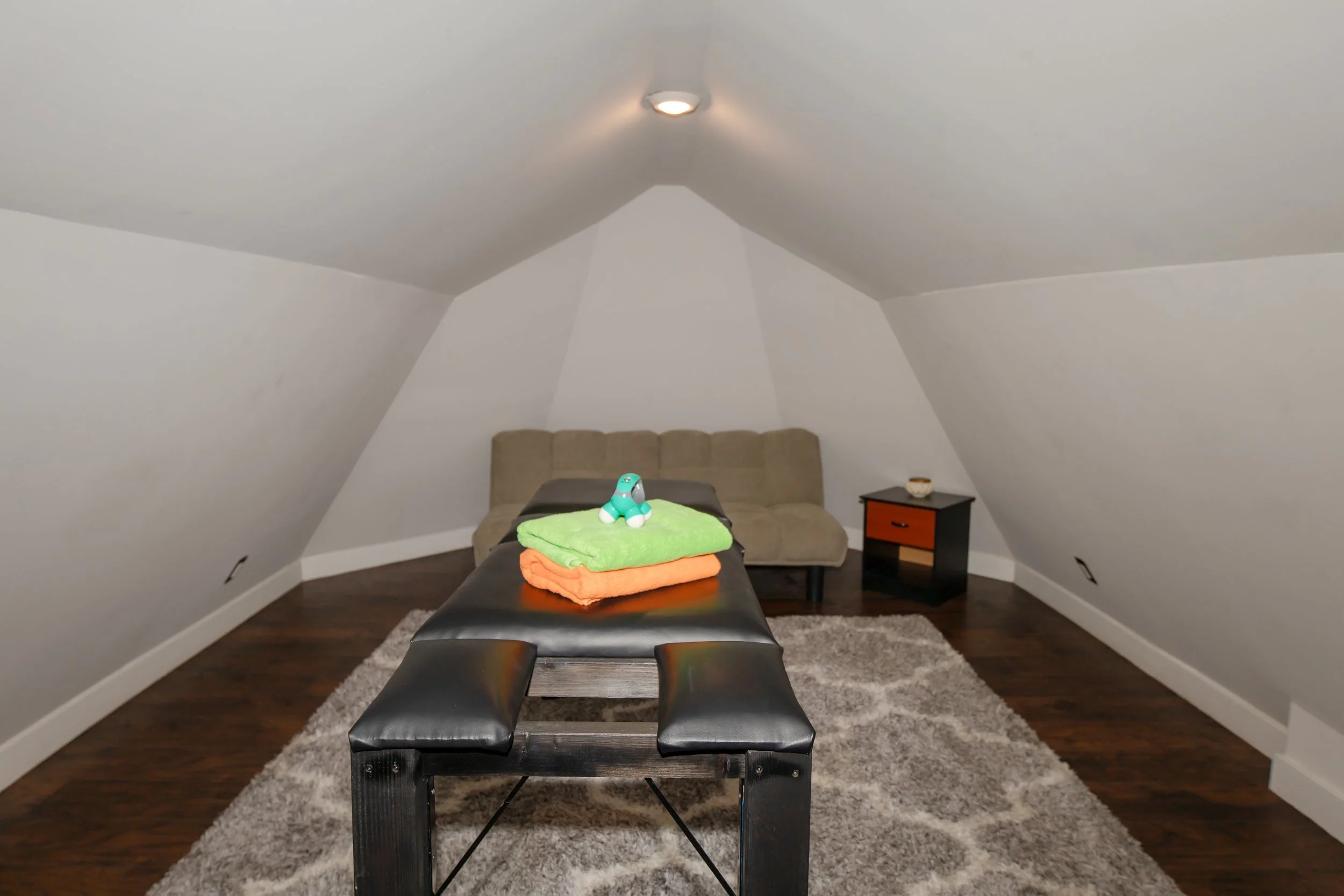 Attic room with sloped ceilings, beige futon against back wall, small black and brown side table, massage table with folded towels and ceramic dog figurine, area rug, ceiling light.