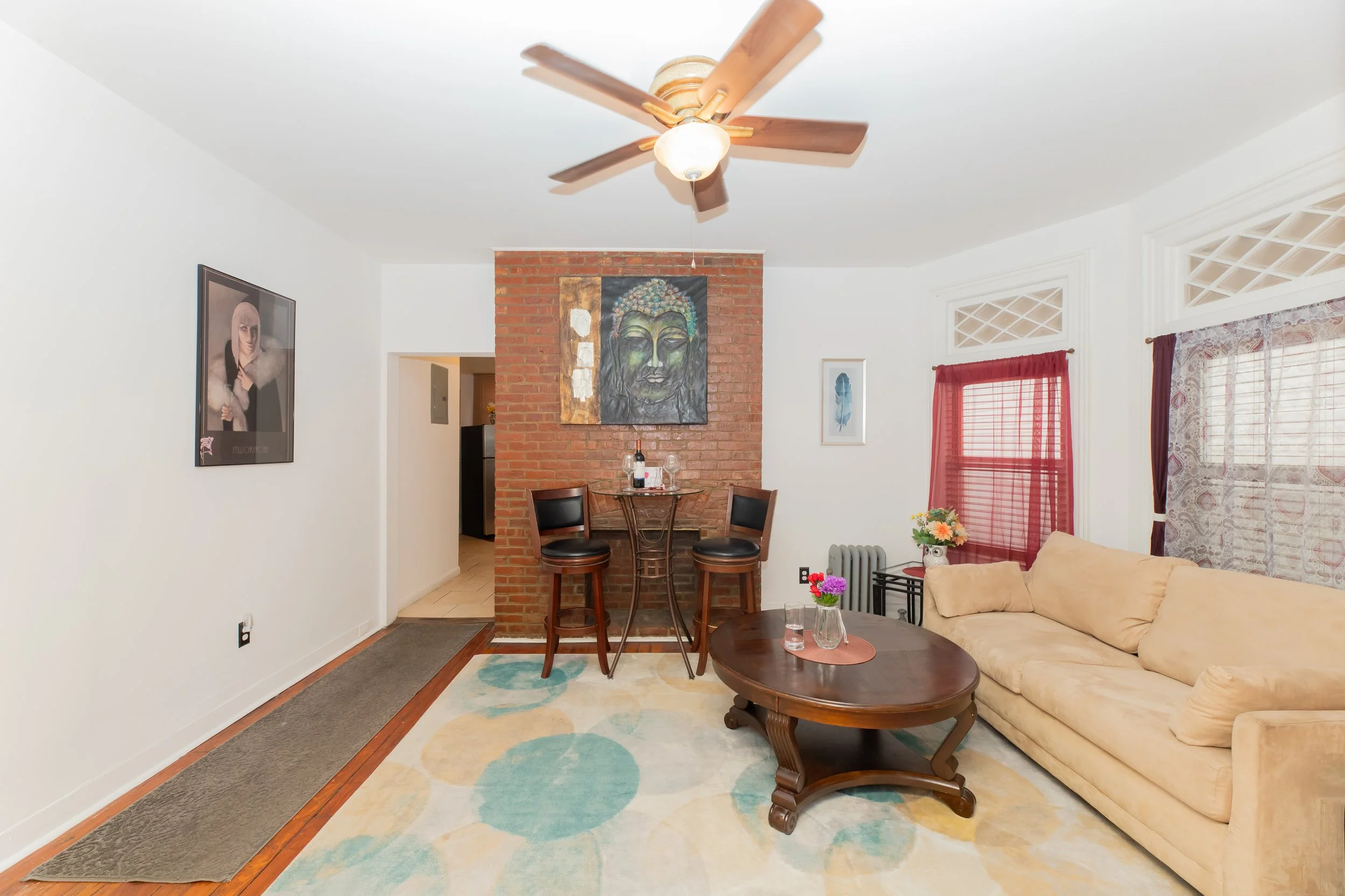 Living room with a yellow sofa, a wooden coffee table with flowers, a beige and red window curtain, a brick wall with a painted Buddha head, a poster of a woman on the white wall, a ceiling fan, and a small bar area with two black chairs.
