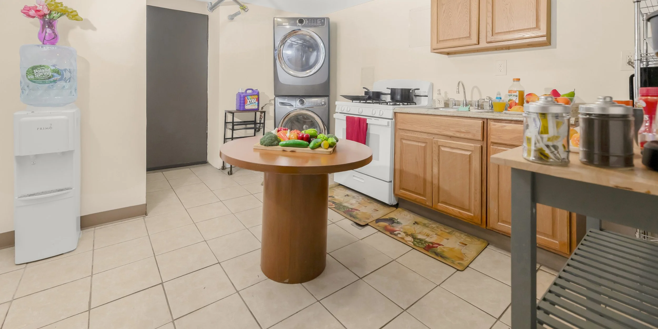 Kitchen with wooden cabinets, a white stove, a dryer and washing machine stacked, a water dispenser with a water jug, and a round wooden table with vegetables on it. There are various kitchen items on counters and shelves, including jars and bottles, as well as a floral rug and tile flooring.