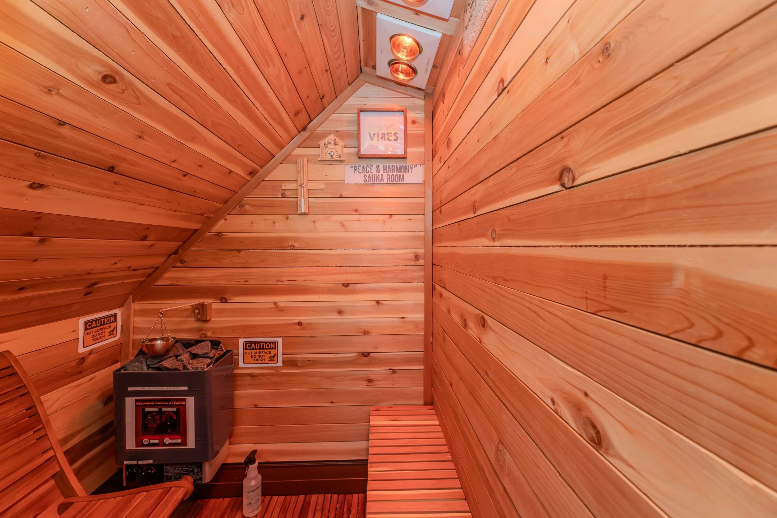Small sauna room with wood-paneled walls and ceiling, sauna heater with stones, wooden bench, and signs indicating caution and sauna room details.