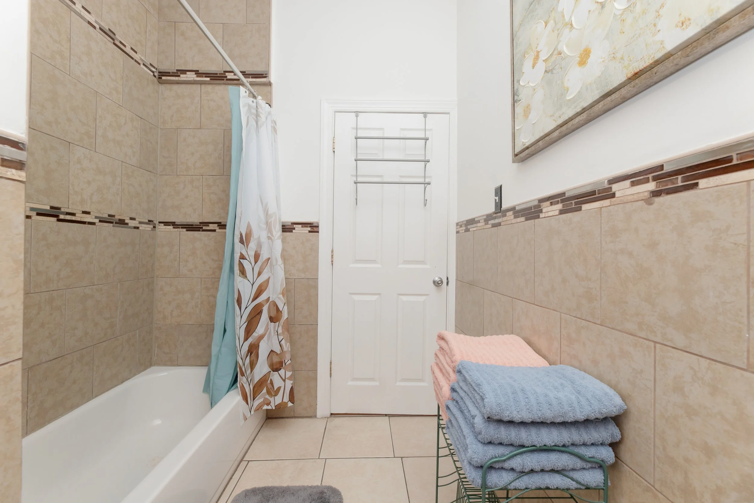 Bathroom with a bathtub, beige tiled walls, a white door, a rack, and stacked towels in pinks and blues.