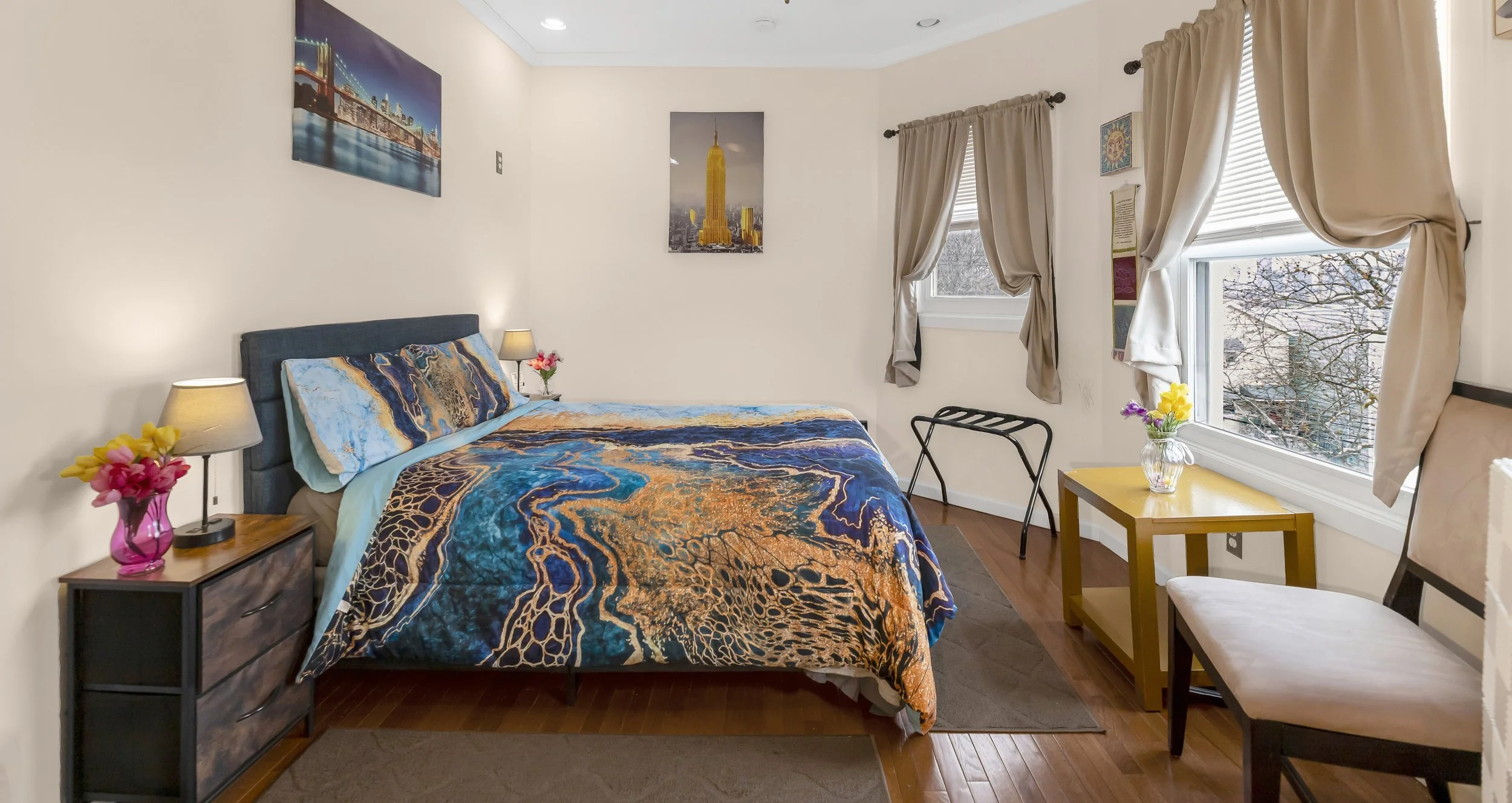 A bedroom with a bed covered in a blue, gold, and black patterned comforter, flanked by matching nightstands with lamps and flowers. The room features beige curtains, two windows, and wall art including cityscape pictures. There is a yellow table with a vase and flowers, a chair, and a luggage rack.
