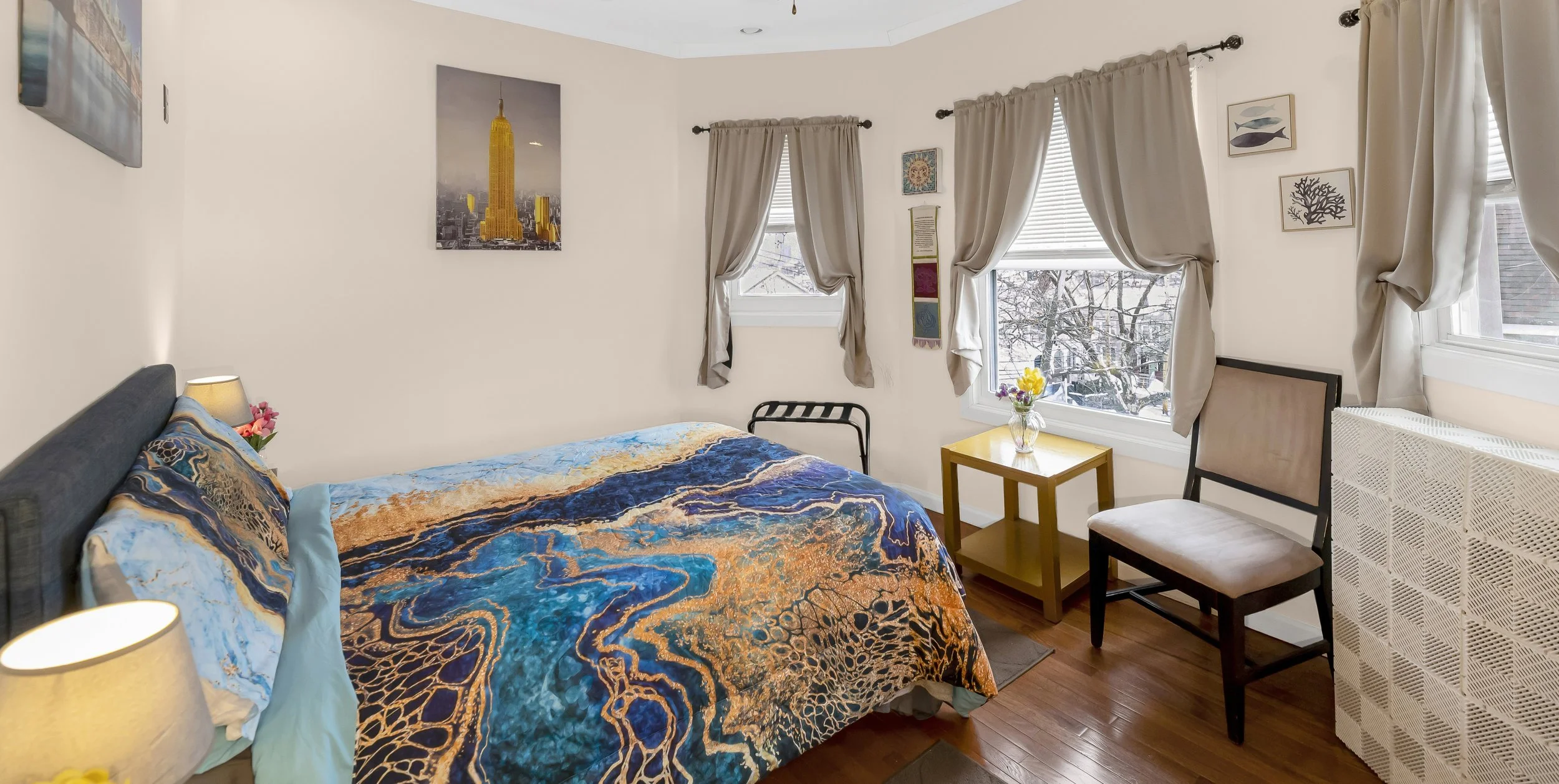 A bedroom with white walls, four windows with beige curtains, a bed with colorful abstract bedding, a yellow side table with flowers, two chairs, and wall art including pictures of fish and trees.