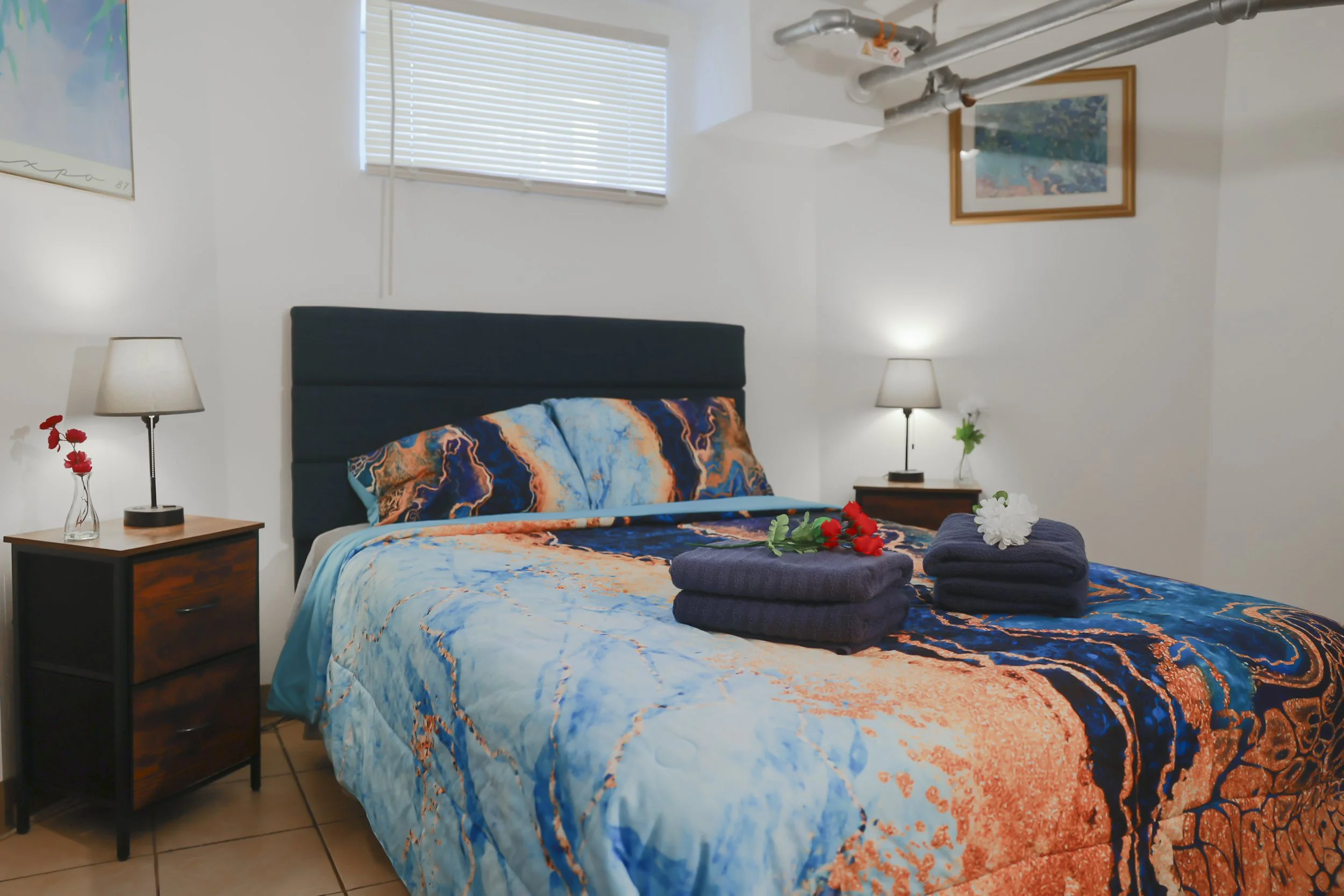 A neatly made bedroom with a bed featuring a vibrant, abstract-colored bedspread and pillows. Two nightstands with lamps and a small vase with flowers are on either side of the bed. Towels and flowers are placed on the bed. There are two wall paintings and a window with blinds, with visible pipes on the ceiling.