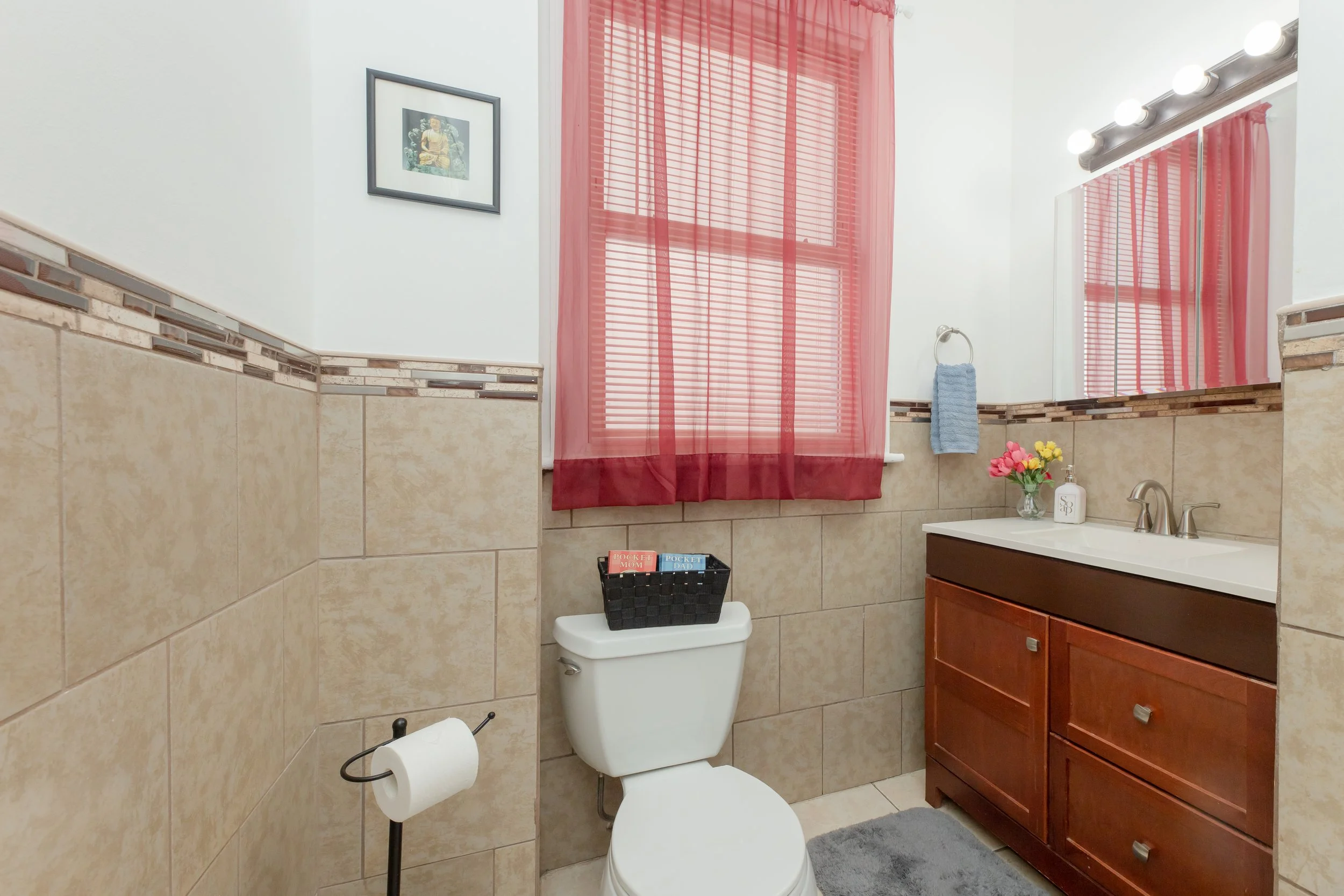 A small bathroom with beige tiled walls, a wooden vanity with a white countertop, a mirror with lights, a window with pink curtains, a framed picture, a blue hand towel, a toilet with a basket of magazines on top, a small rug, and a vase of flowers on the sink.