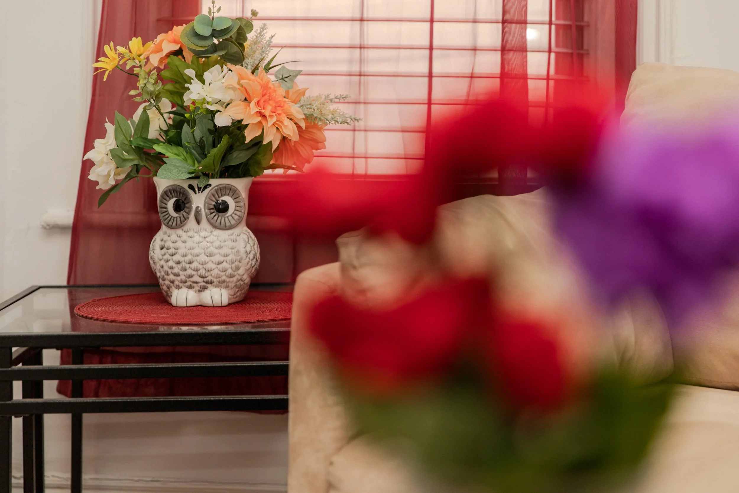 A decorative owl-shaped vase with large eyes holding a bouquet of peach, white, and yellow flowers and green foliage, placed on a red mat on a glass-topped table near a window with red curtains in a cozy living room.