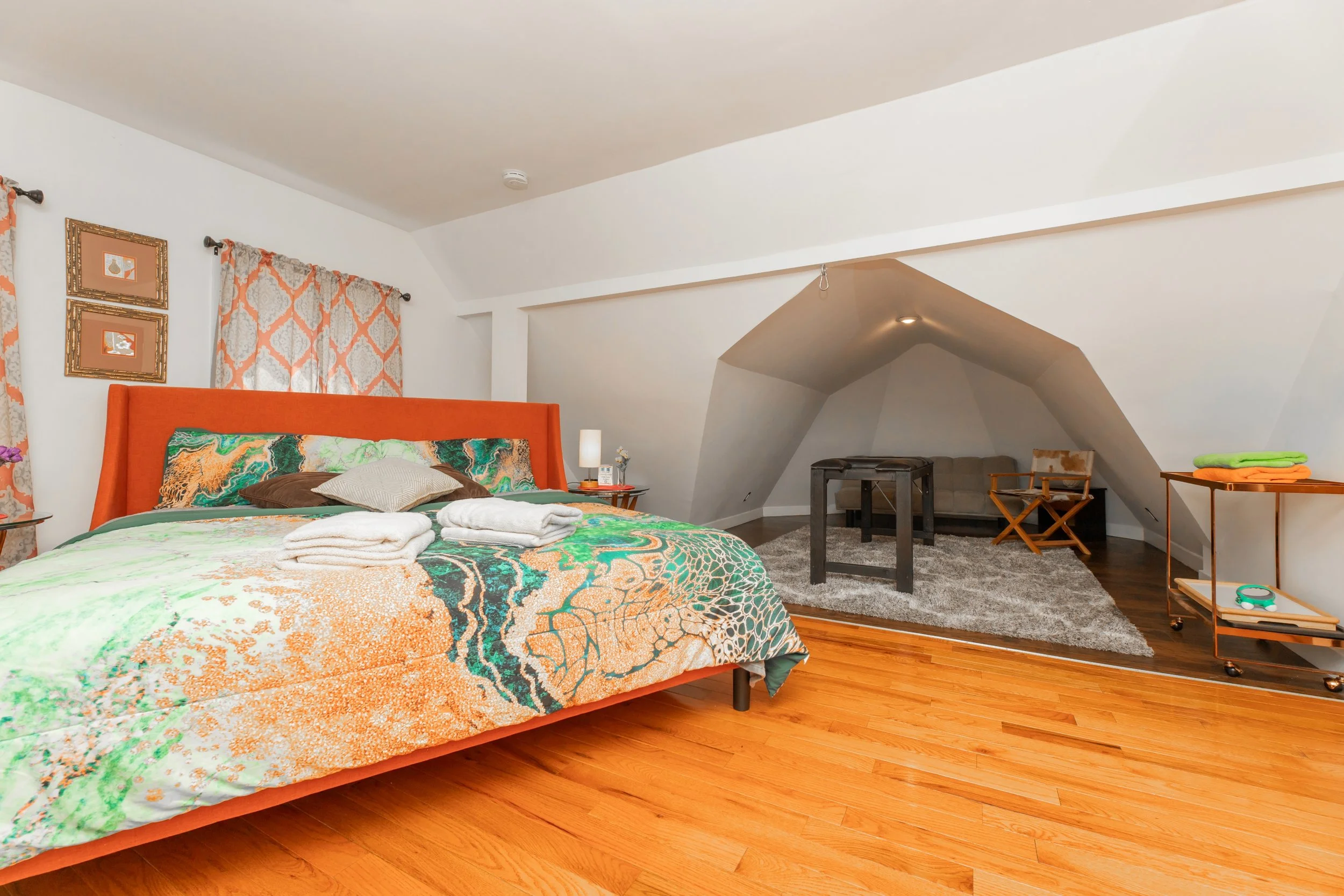 A cozy bedroom with a large bed featuring an orange headboard, colorful patterned bedding, and folded towels. The room has hardwood floors, decorative curtains, and artwork on the wall. There's a small sitting area with a gray rug, a sofa, and a chair in a nook with sloped ceiling, along with a side table and cart with towels and a timer.