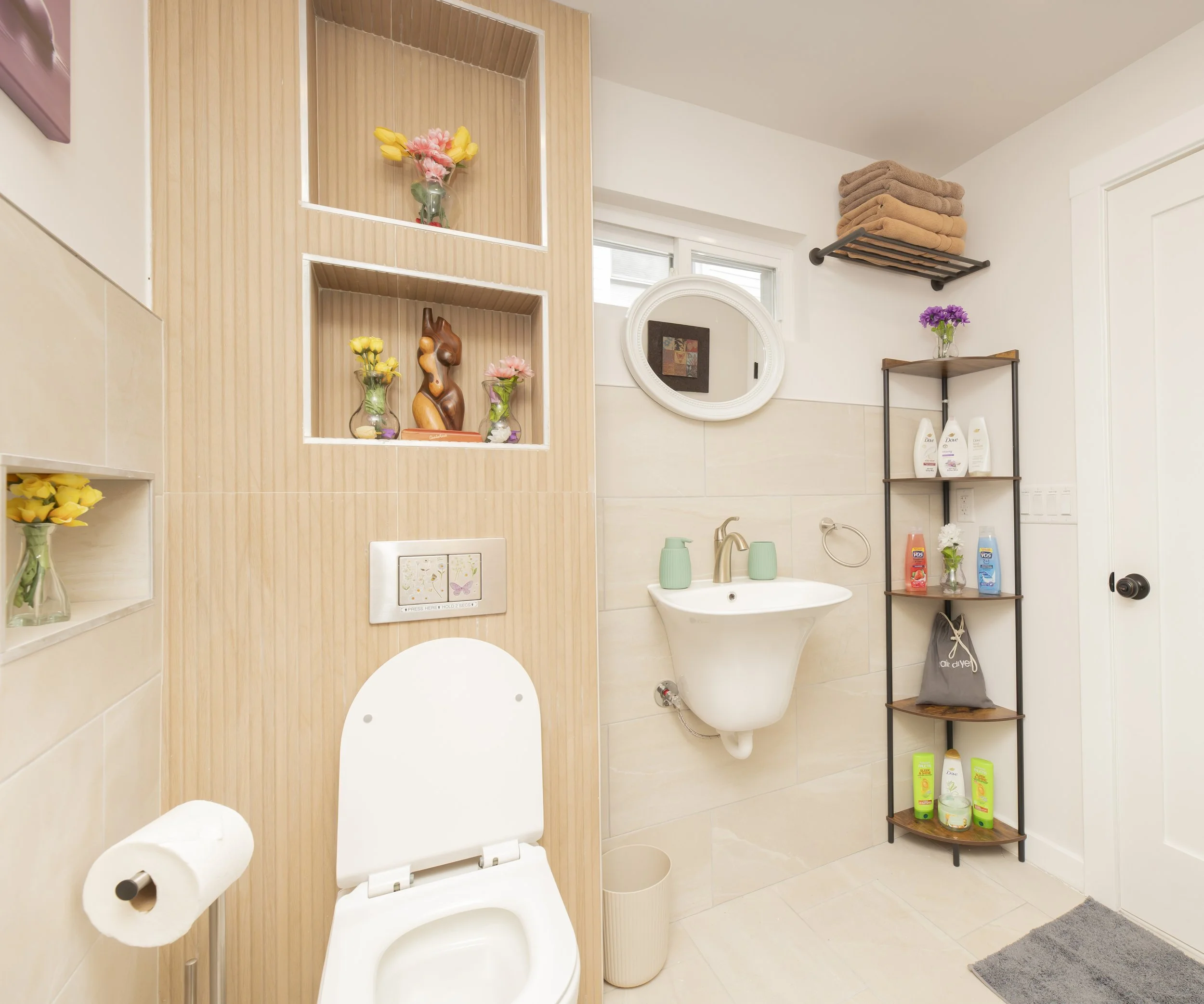 A small bathroom with a wall-mounted toilet, a white sink, and a black shelving unit holding toiletries and towels. Decorative vases with flowers are on shelves and surfaces, with a small window and a round mirror above the sink.