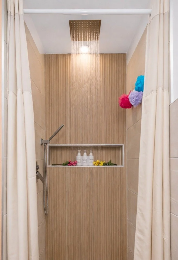 A modern shower with a ceiling rain showerhead, beige tiled walls, and a small shelf holding three bottles of shower products and colorful flowers. Cream curtains frame the shower.