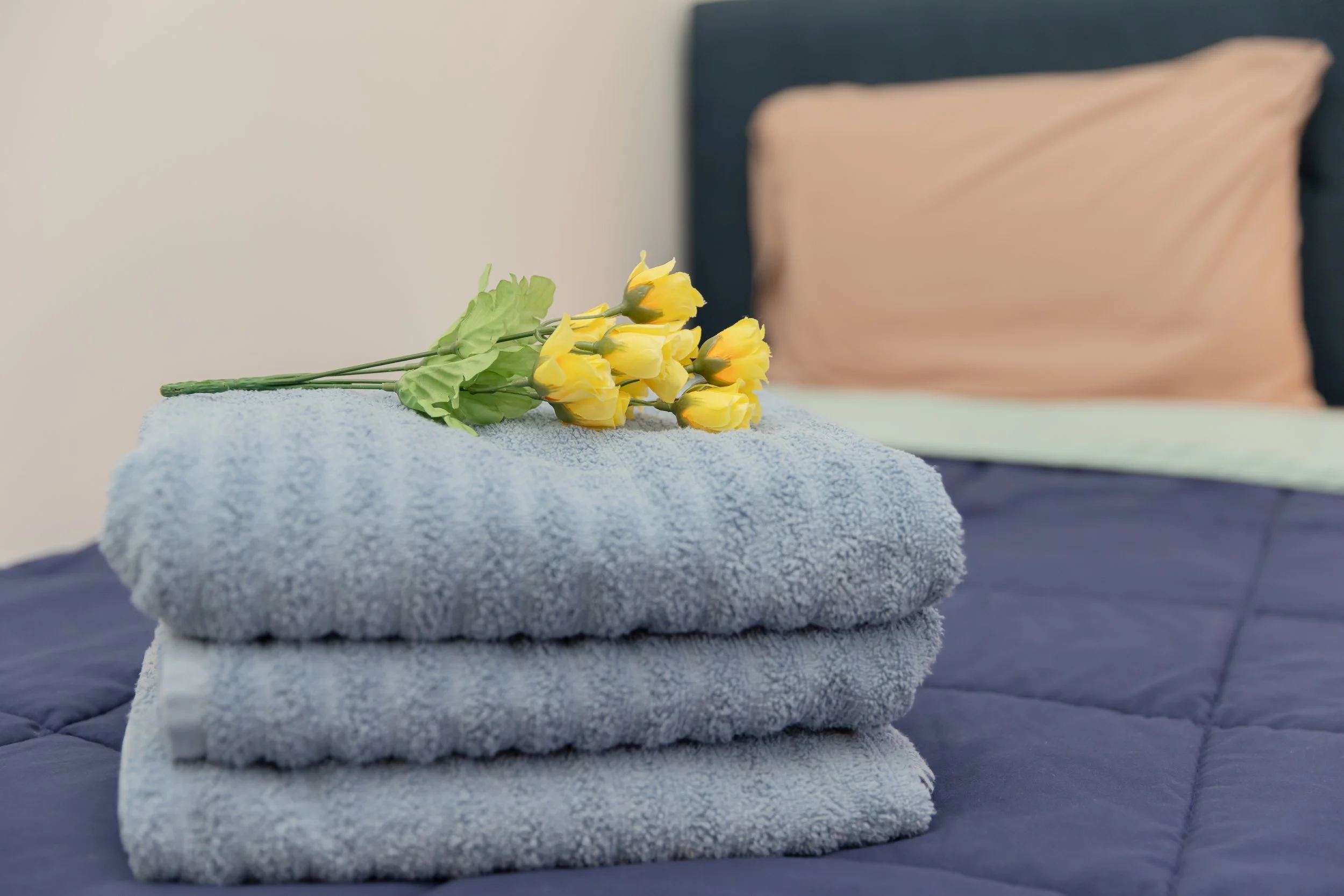 A stack of folded blue towels on a bed with a bouquet of yellow flowers on top, and a pink pillow in the background