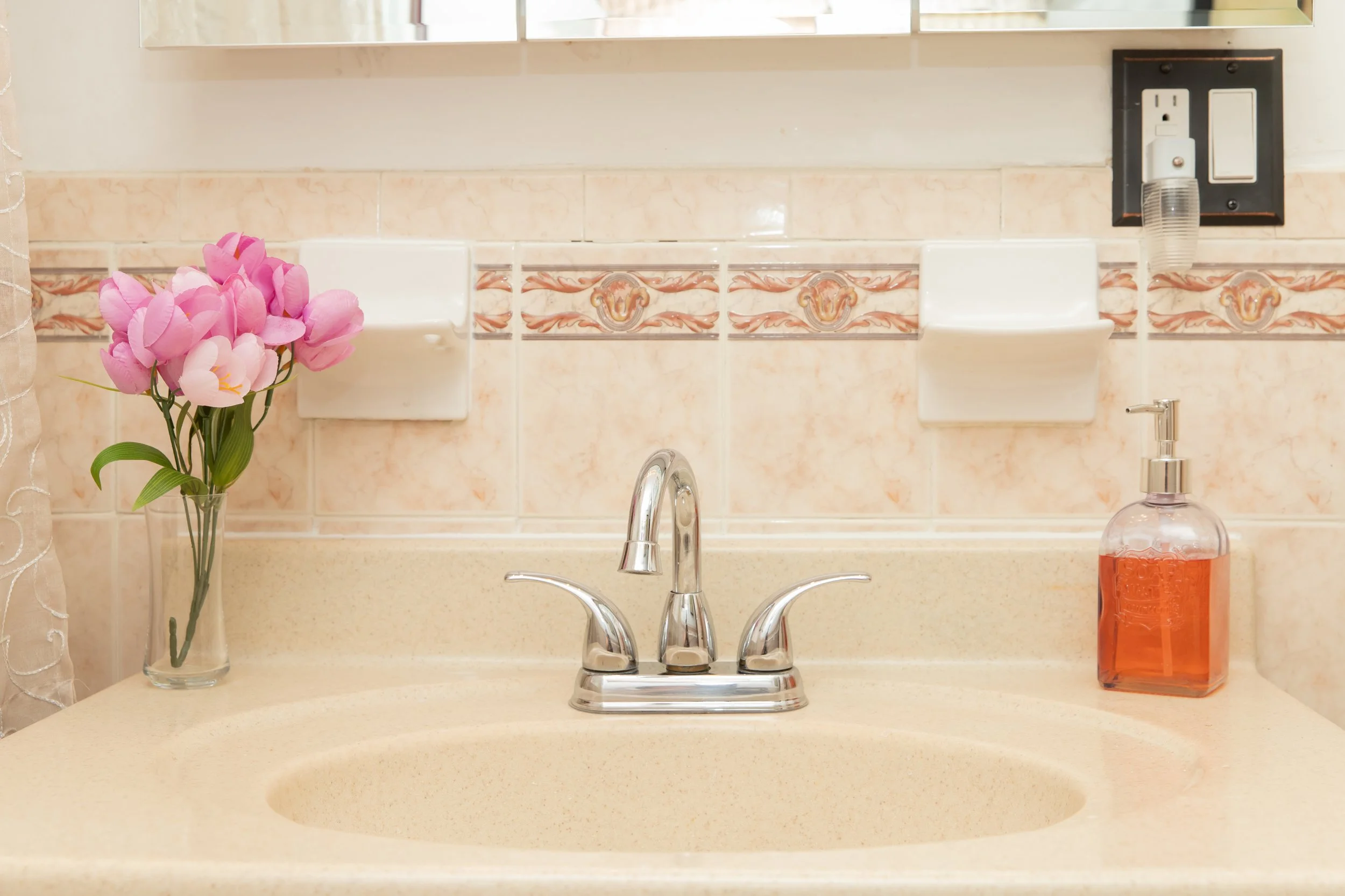 Bathroom sink area with pink artificial flower bouquet on the left, a soap dispenser with orange liquid on the right, a silver faucet in the center, and a mirror with a power outlet and cover above.