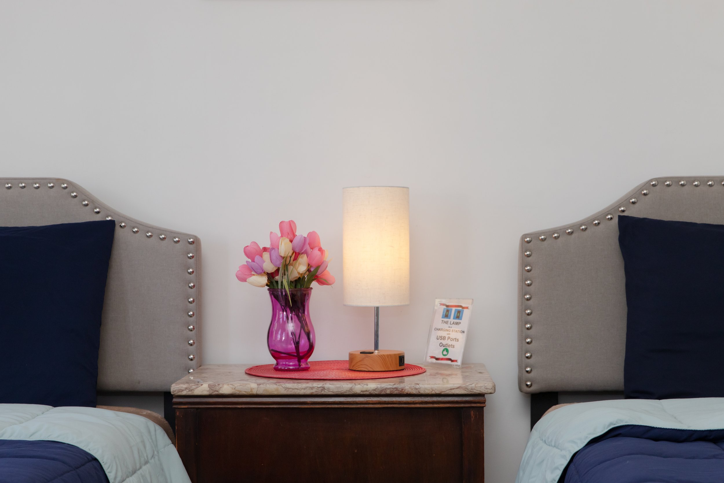 Decorative bedroom scene with two beds, a nightstand, a table lamp, a vase of pink and white tulips, and a small informational sign.