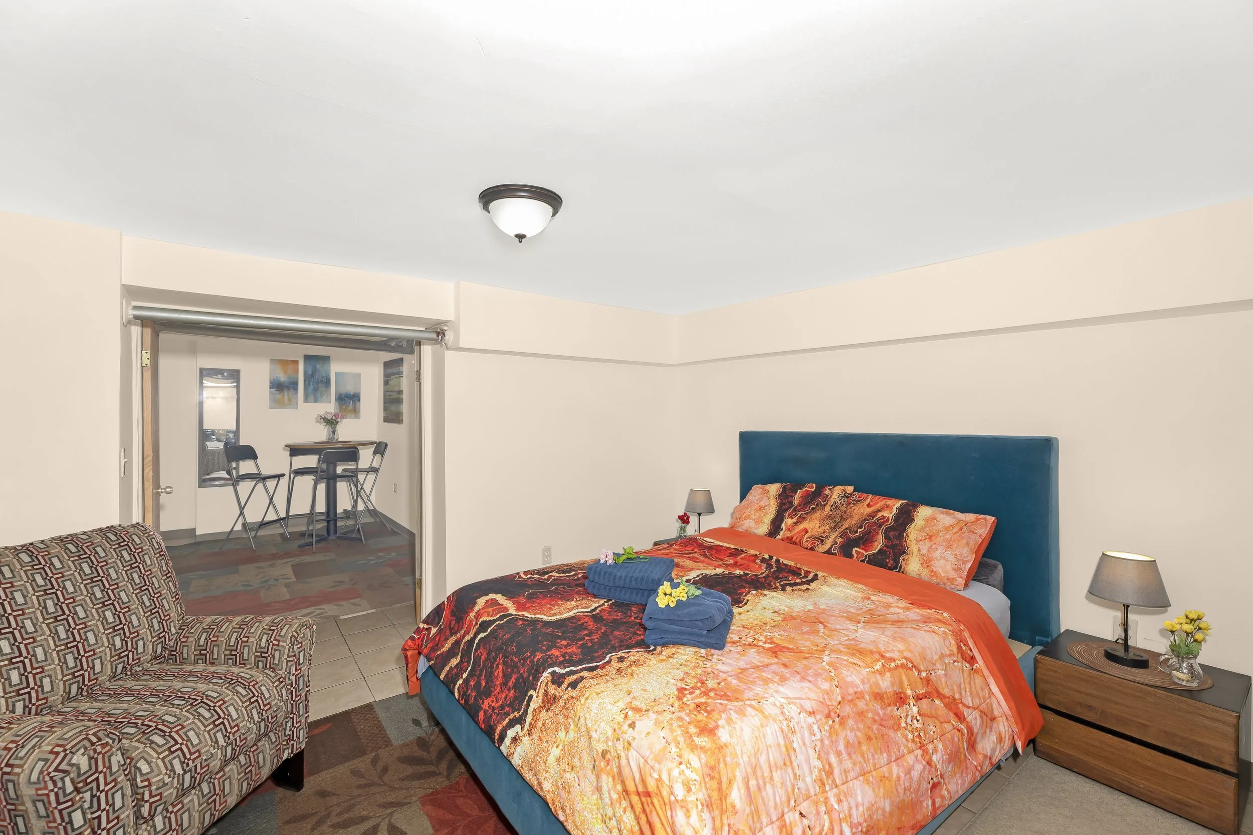 A bedroom with a bed that has a colorful orange, red, black, and yellow duvet and a teal headboard. There are two folded blue towels with flowers on them on the bed. To the right of the bed, there is a wooden nightstand with a gray lamp, a small vase with yellow flowers, and a remote control. To the left, there's a patterned armchair. In the background, an opening leads to a small dining area with a high table, three chairs, and wall art. The room has beige walls and tiled flooring.