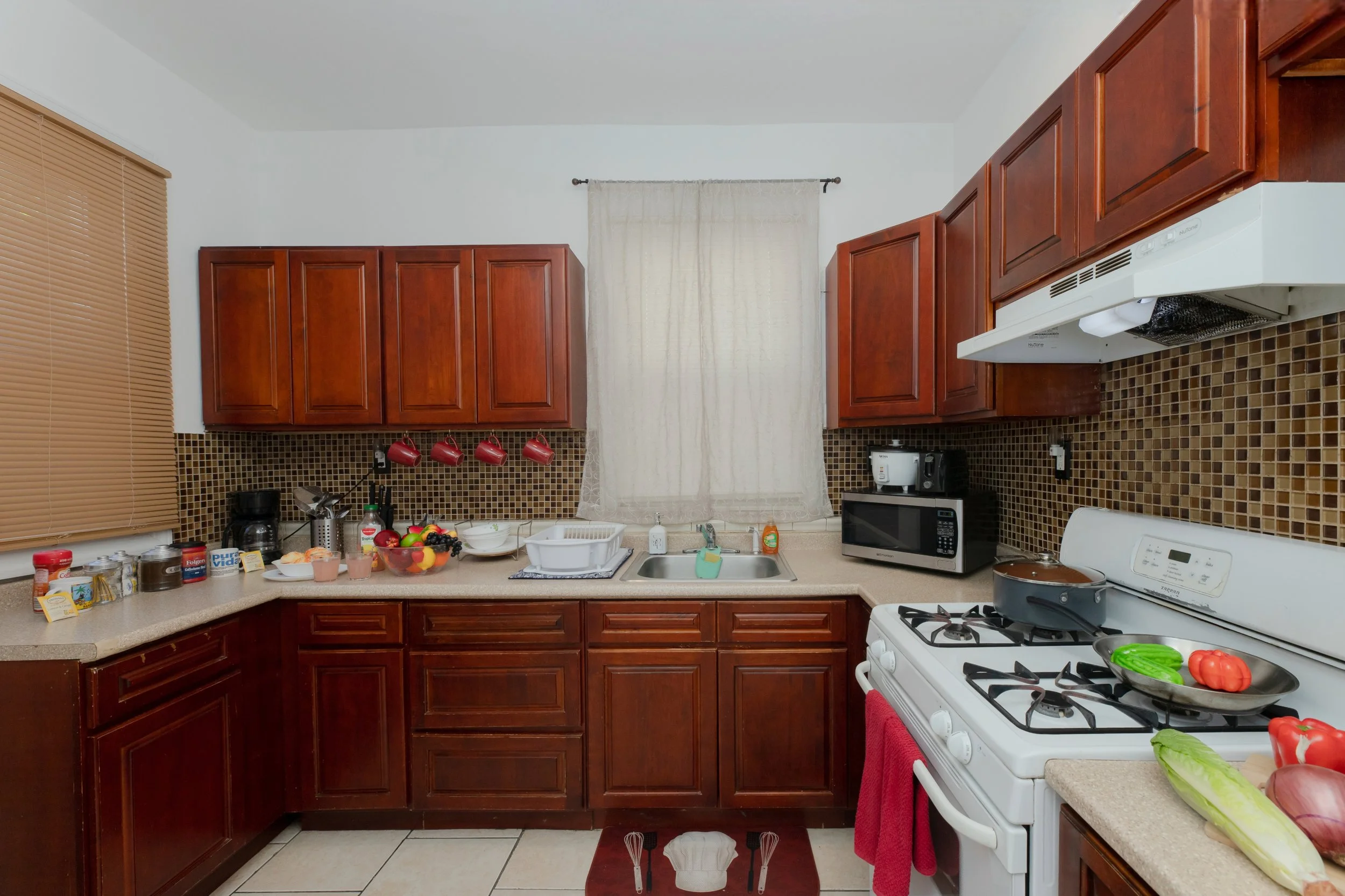 Kitchen with wooden cabinets, beige countertops, a white stove, microwave, toaster, and coffee machine. Contains fresh vegetables, fruit, and kitchen utensils.