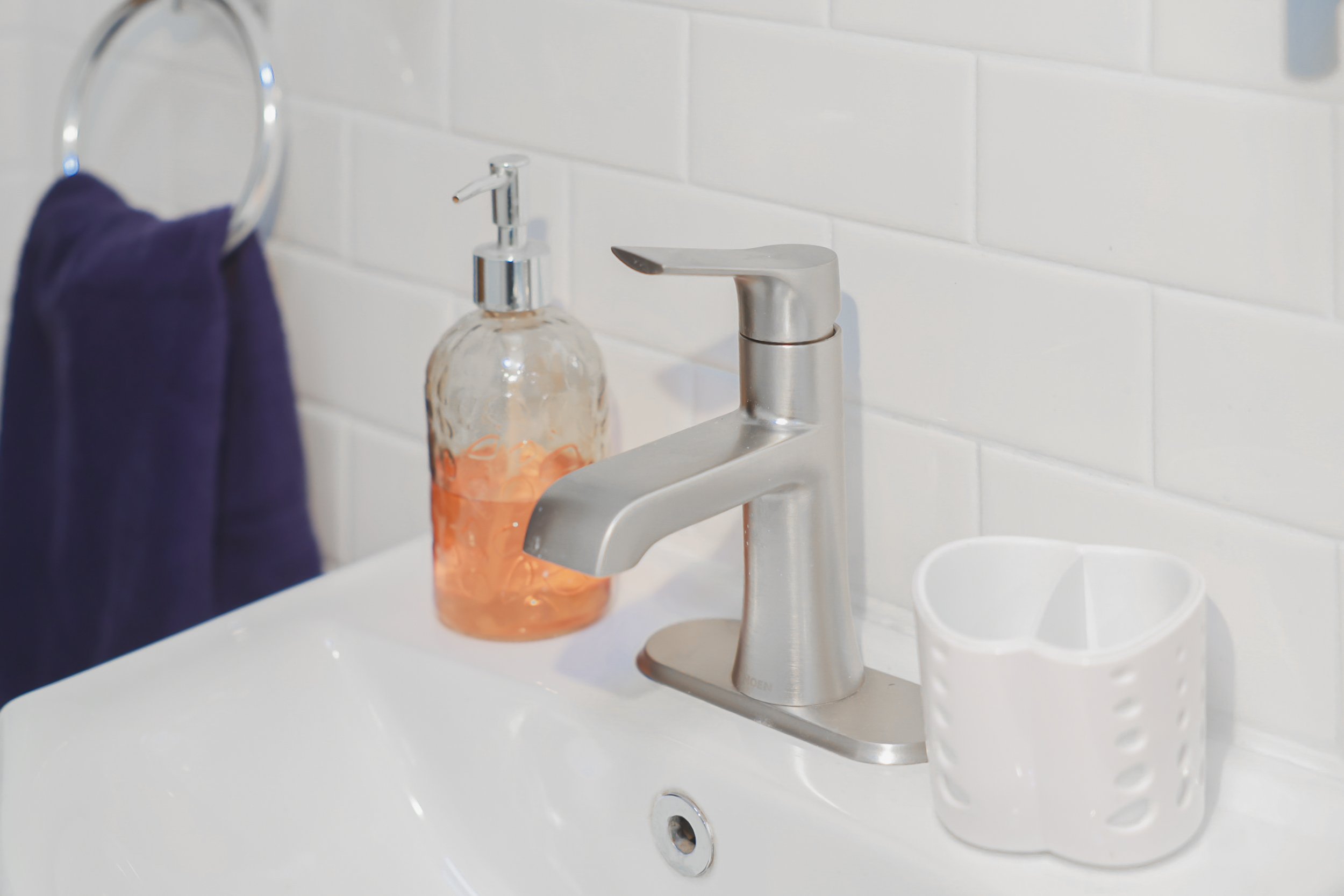 Bathroom sink with a silver faucet, a clear soap dispenser with orange liquid soap, a white toothbrush holder, and a purple towel hanging on a ring, against white tiled wall.