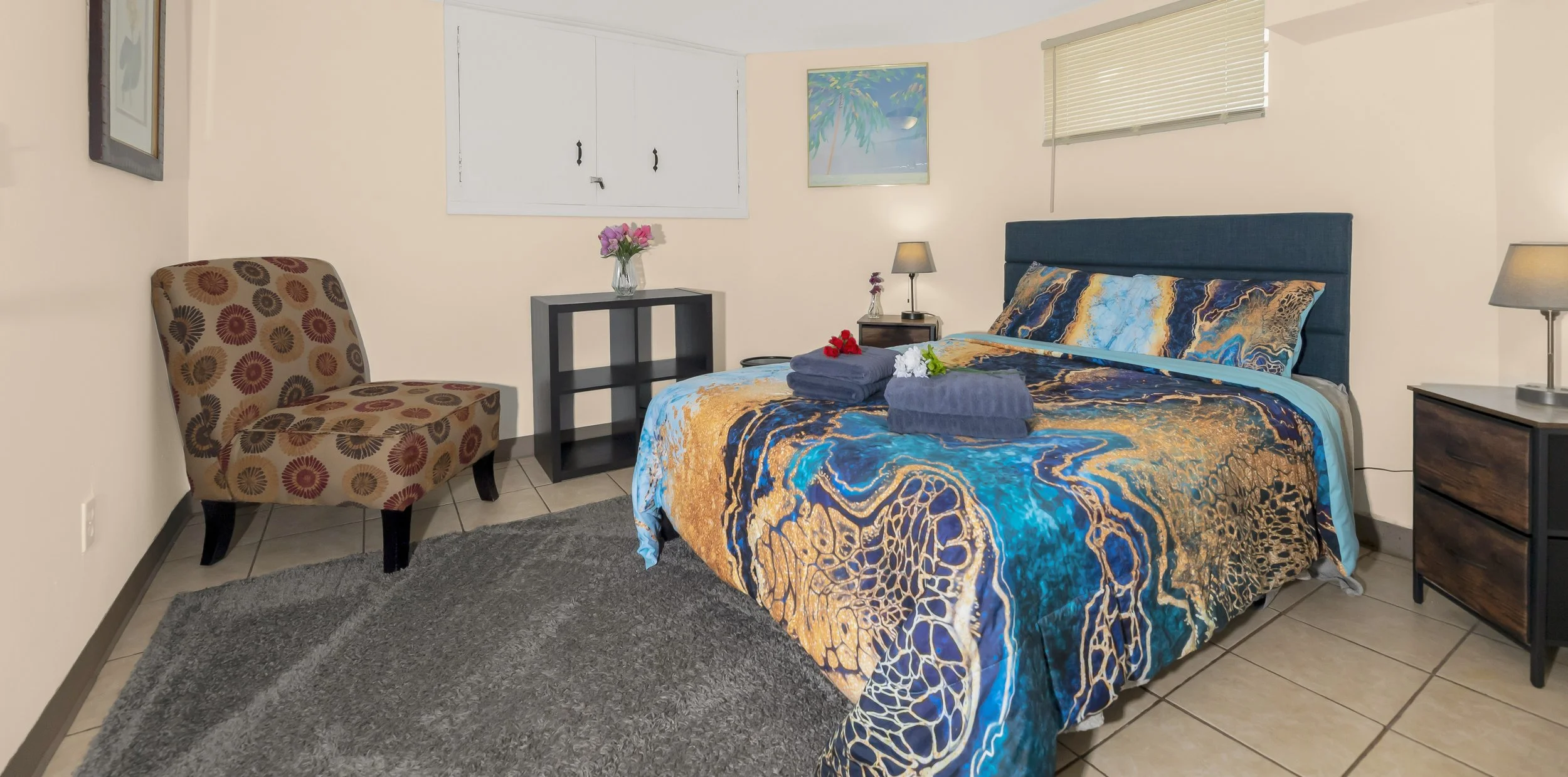 A cozy bedroom with a neatly made bed featuring a colorful blue and gold patterned bedspread. On the bed are two folded towels with small flowers. There's a nightstand with a lamp, a wall art of a beach scene, a chair with circular patterns, a small black shelf with a vase of pink flowers, and a window with blinds. The room has beige walls, tile flooring, and a soft gray rug.