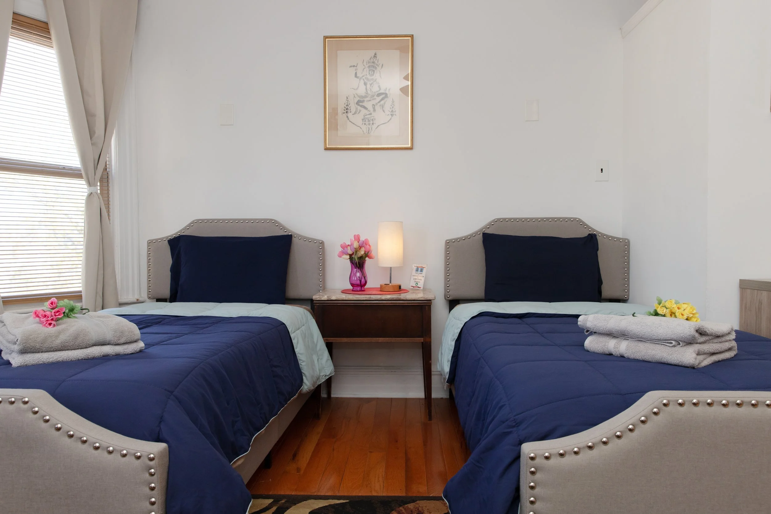 A bedroom with two twin beds with beige upholstered headboards and blue bedding. Each bed has a rolled towel with flowers on top. A wooden nightstand between the beds holds a pink vase with pink flowers, a table lamp, and a small table card. There is a window with blinds and curtains to the left, and a framed artwork on the white wall above the nightstand.