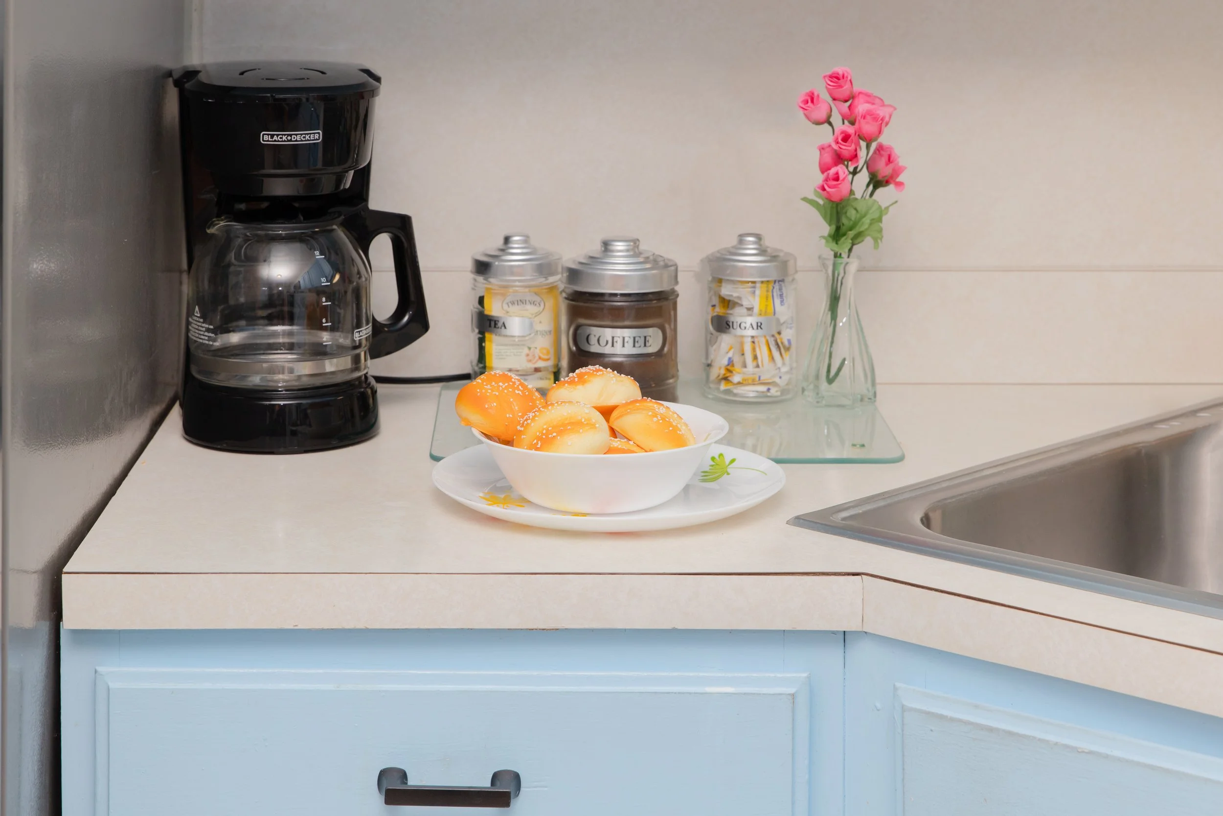 Coffee maker, jars labeled tea, coffee, sugar, a bowl of bread rolls, and a vase with pink artificial flowers on a kitchen countertop.