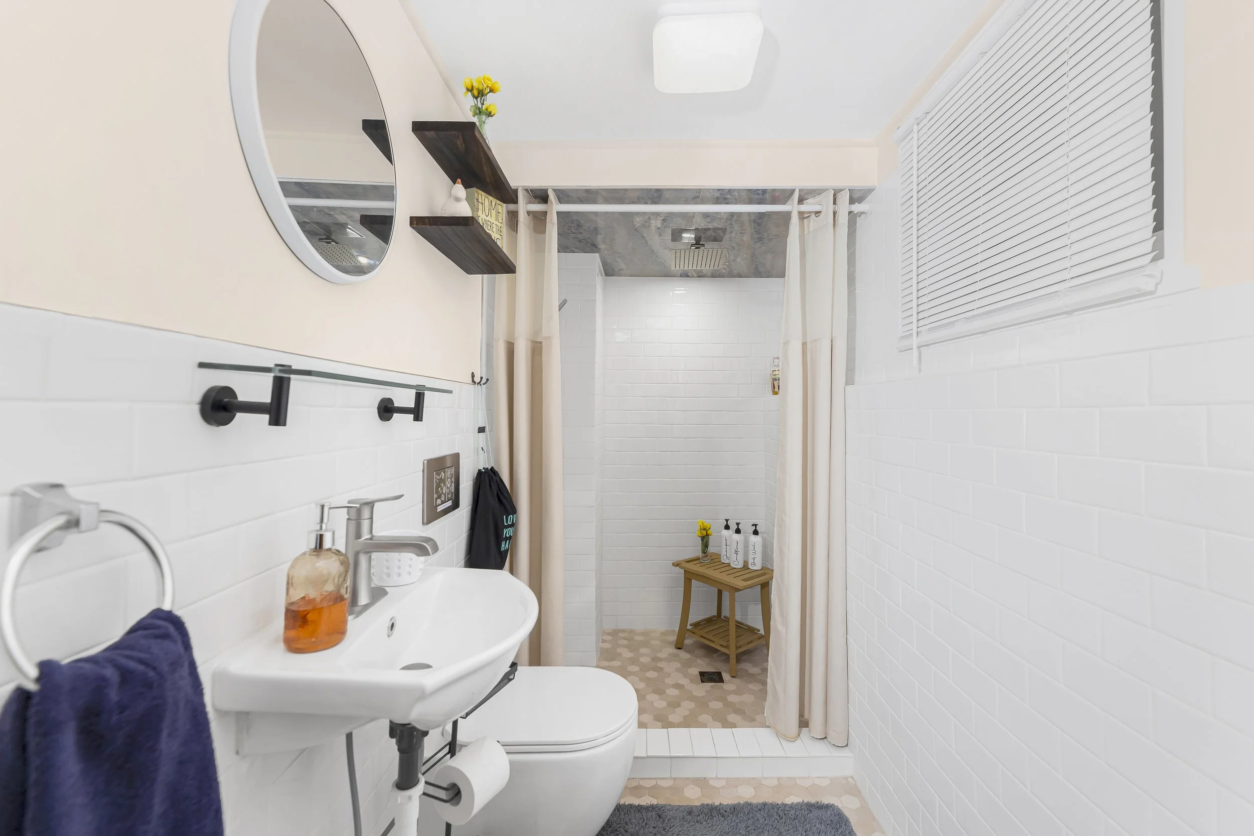 A small bathroom with a white brick wall and patchy pink hexagon tile floor, featuring a shower area with beige curtains, a small wooden bench, and a window with white blinds on the right side. There is a white ceramic toilet with a small hand-wash basin, a navy towel, a soap dispenser, and a mirror on the beige wall, and two dark brown wall-mounted shelves with a plant and decorative items.
