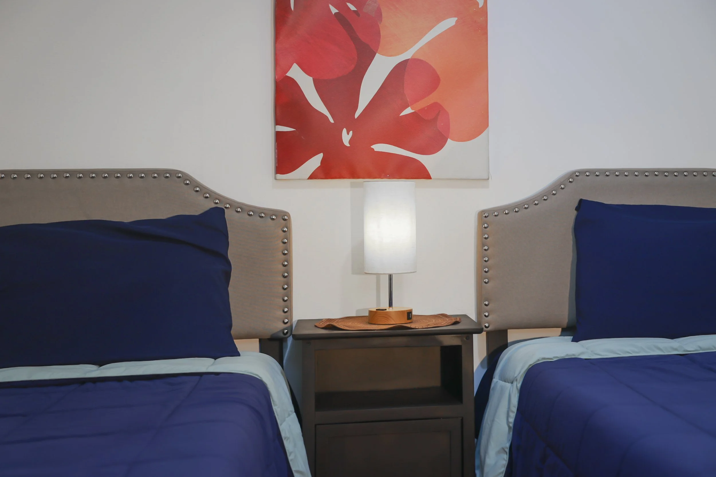 A hotel room with two beds, each with dark blue bedding and pillows, separated by a nightstand with a lamp, and a red-orange floral painting on the wall behind the lamp.