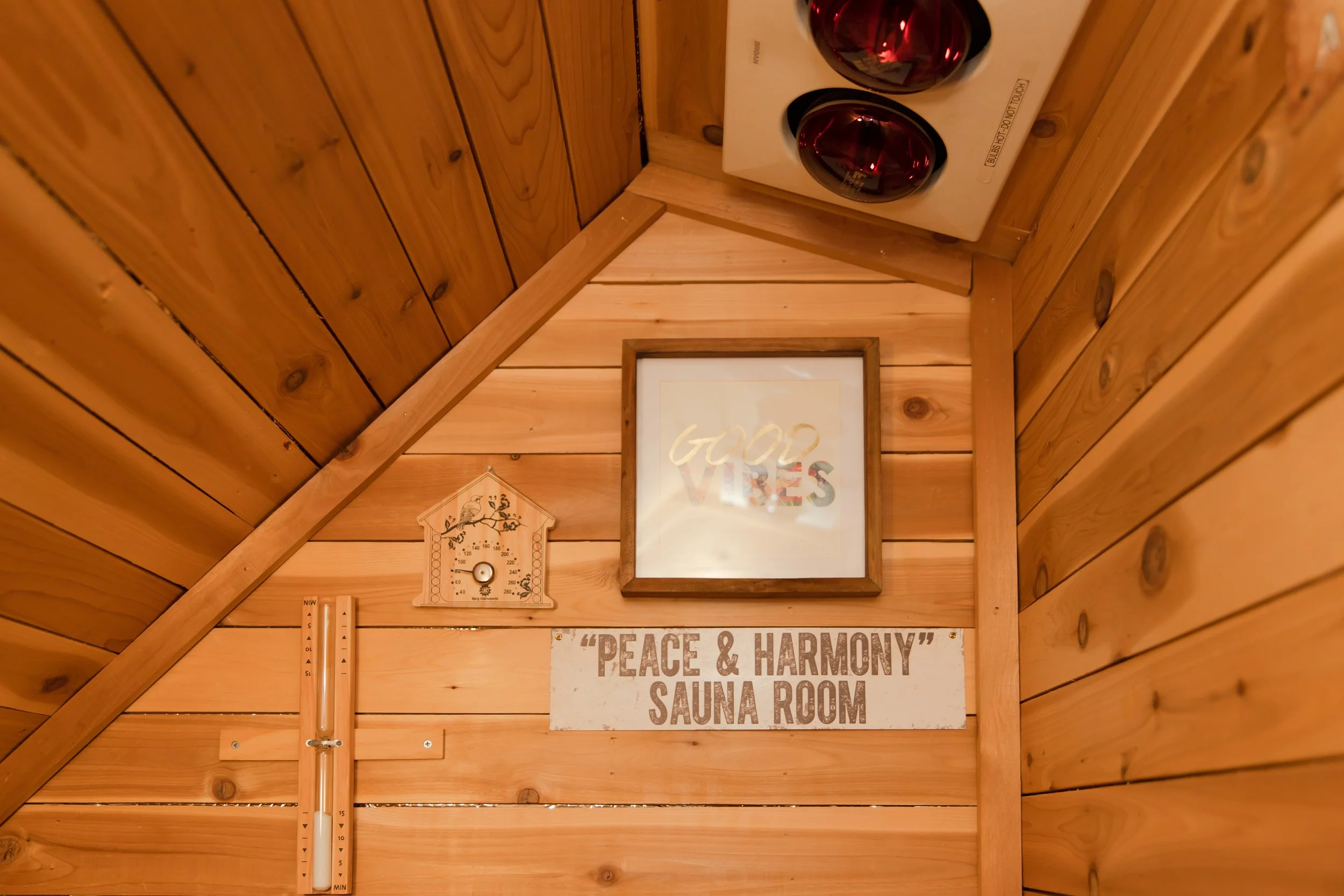 Wood-paneled wall in a sauna room with a sign that reads "Peace & Harmony Sauna Room," a framed quote that says "God Loves," a sauna thermometer, and an electric stove with red stones.