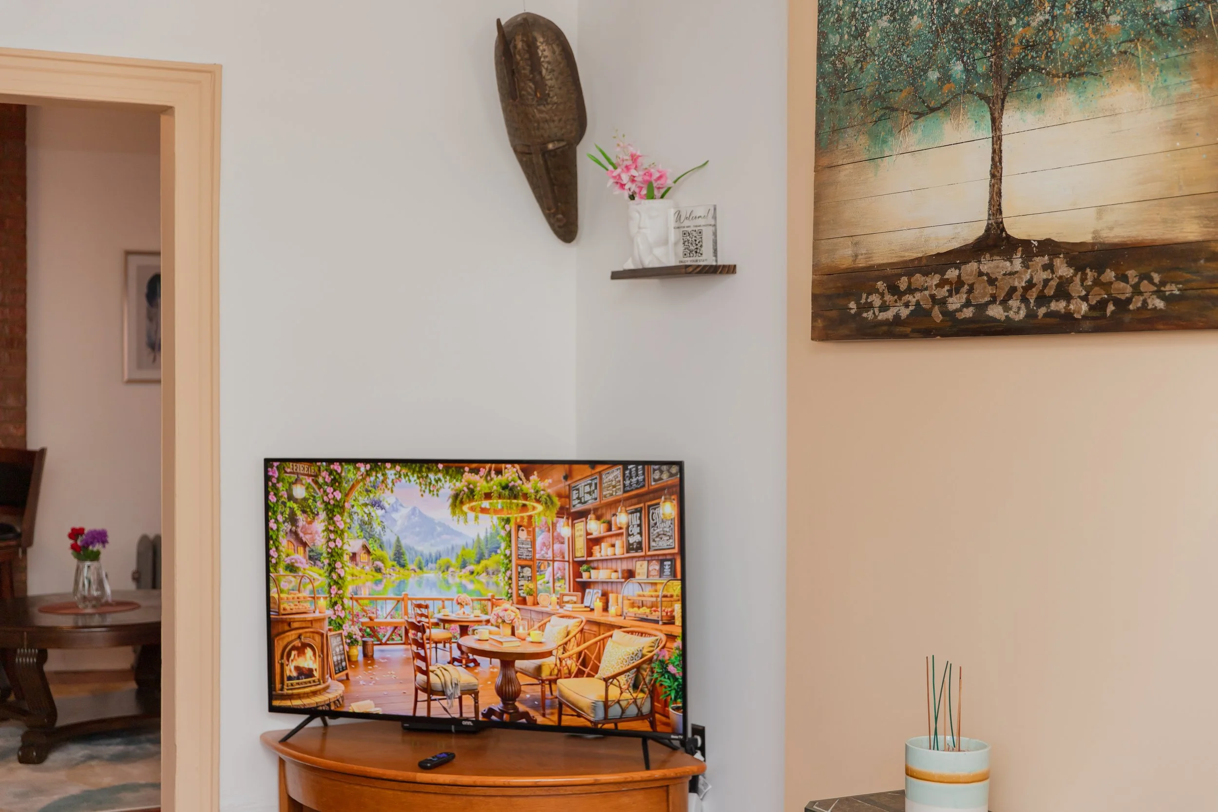 Living room corner with a flat-screen TV on a wooden desk displaying a colorful mountain scene, a remote control in front, a white vase with pink flowers and a QR code on a floating shelf, a tribal mask wall art, and a large painting of a tree, with an adjacent room visible through a doorway.
