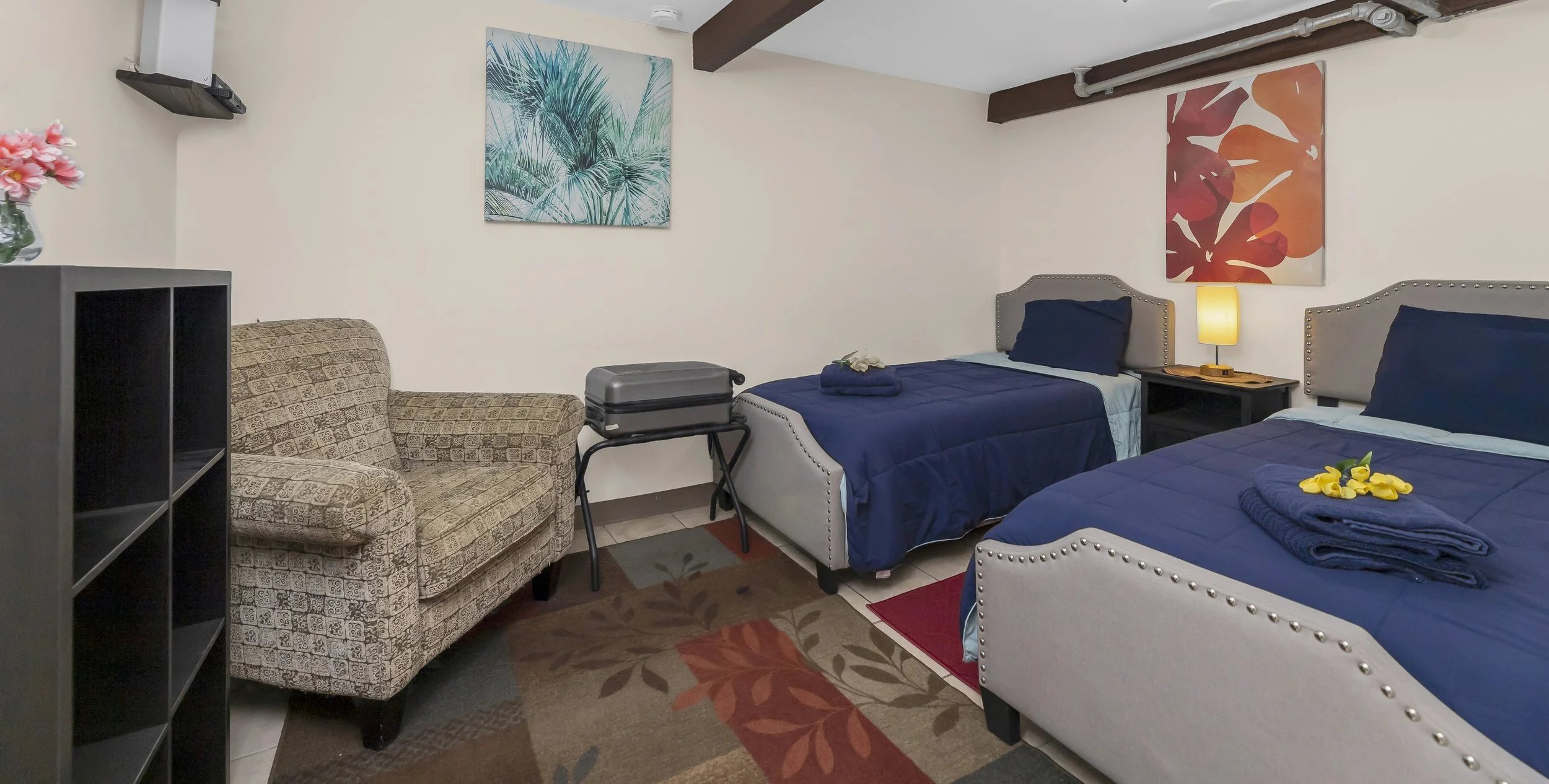 Room with two beds, each with navy blue comforters and navy pillows. A nightstand with a lamp between the beds, and towels with flowers on each bed. A patterned armchair, a small table with a luggage stand, a black bookshelf, and wall art featuring tropical themes.