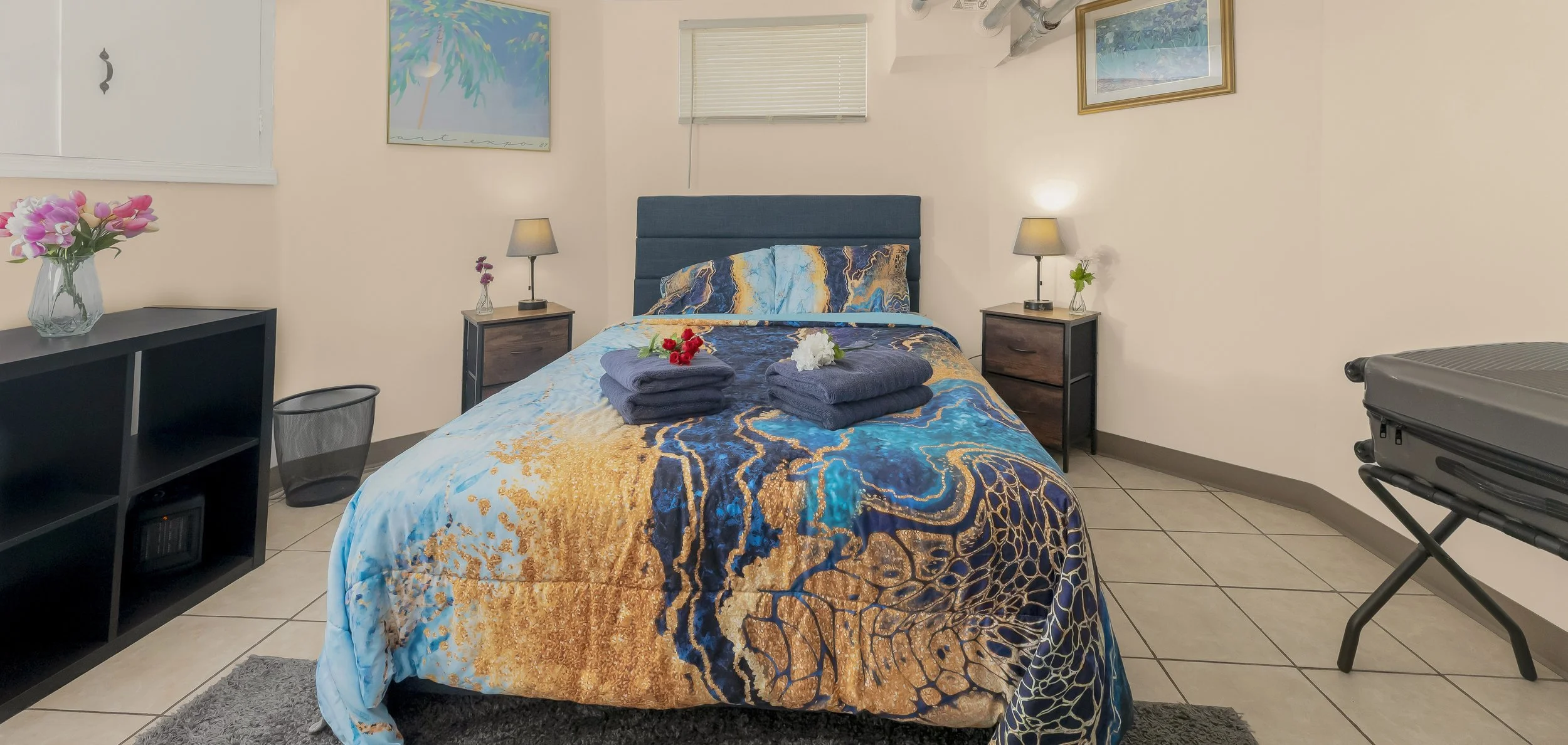 A neatly made bed with a multicolored bedspread featuring blue, gold, and black patterns, and two folded dark gray towels with flowers on top. The bed has a blue upholstered headboard and is flanked by two black nightstands with lamps. There is a window with closed blinds above the bed, framed artwork on the walls, a black bookshelf with a vase of pink flowers on the left, a trash bin below it, and a black suitcase on a foldable stand on the right. The room has tiled flooring and light-colored walls.