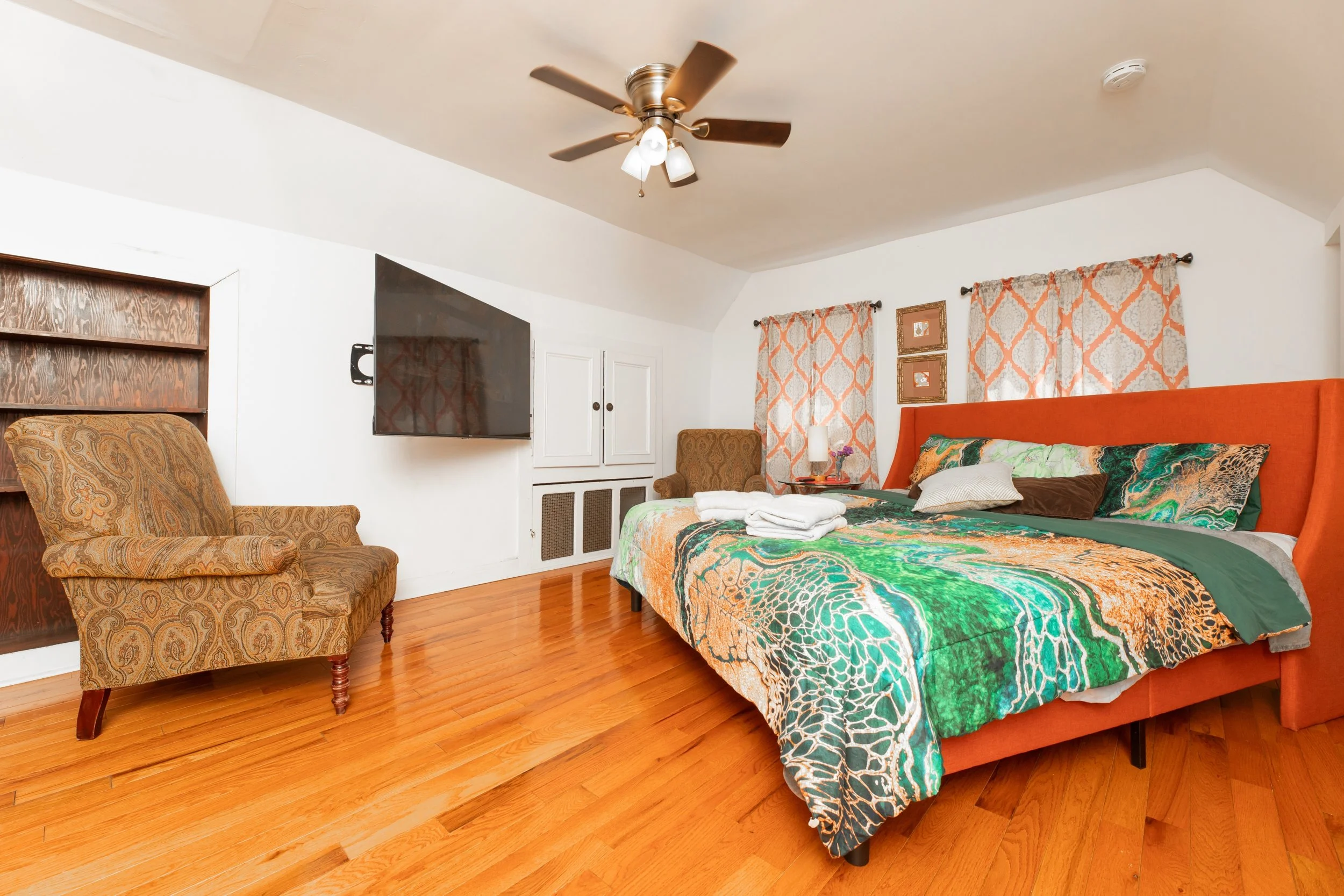 A bedroom with a large bed, colorful bedding, and an orange headboard. There is a patterned armchair, a wall-mounted TV, and a small beige chair near the window with orange and beige curtains. The room has hardwood floors and a ceiling fan with lights.