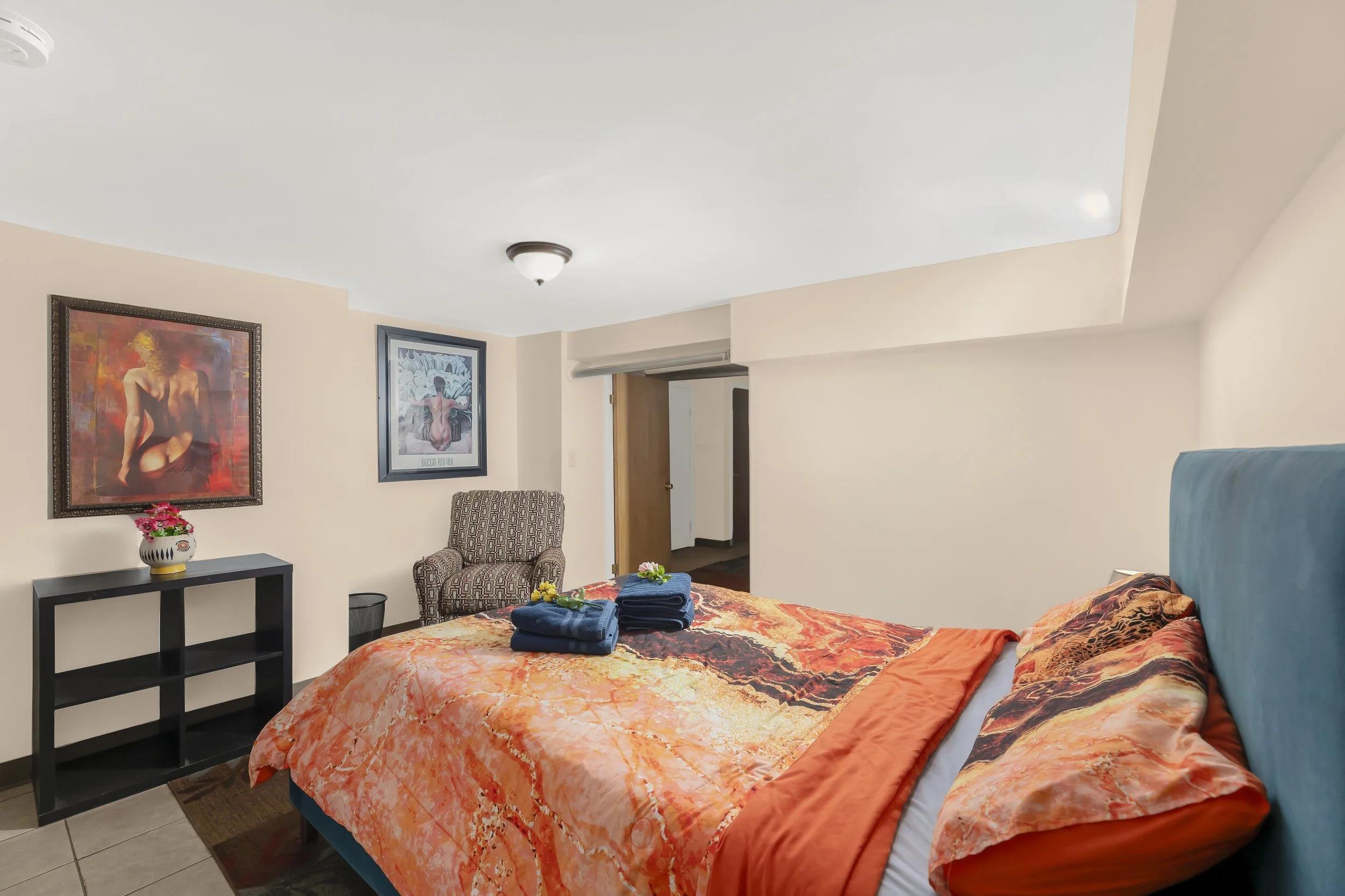 Hotel room with a bed, colorful bedding, and towels. Artworks on the wall, a patterned armchair, and a small black shelf with a flower vase.