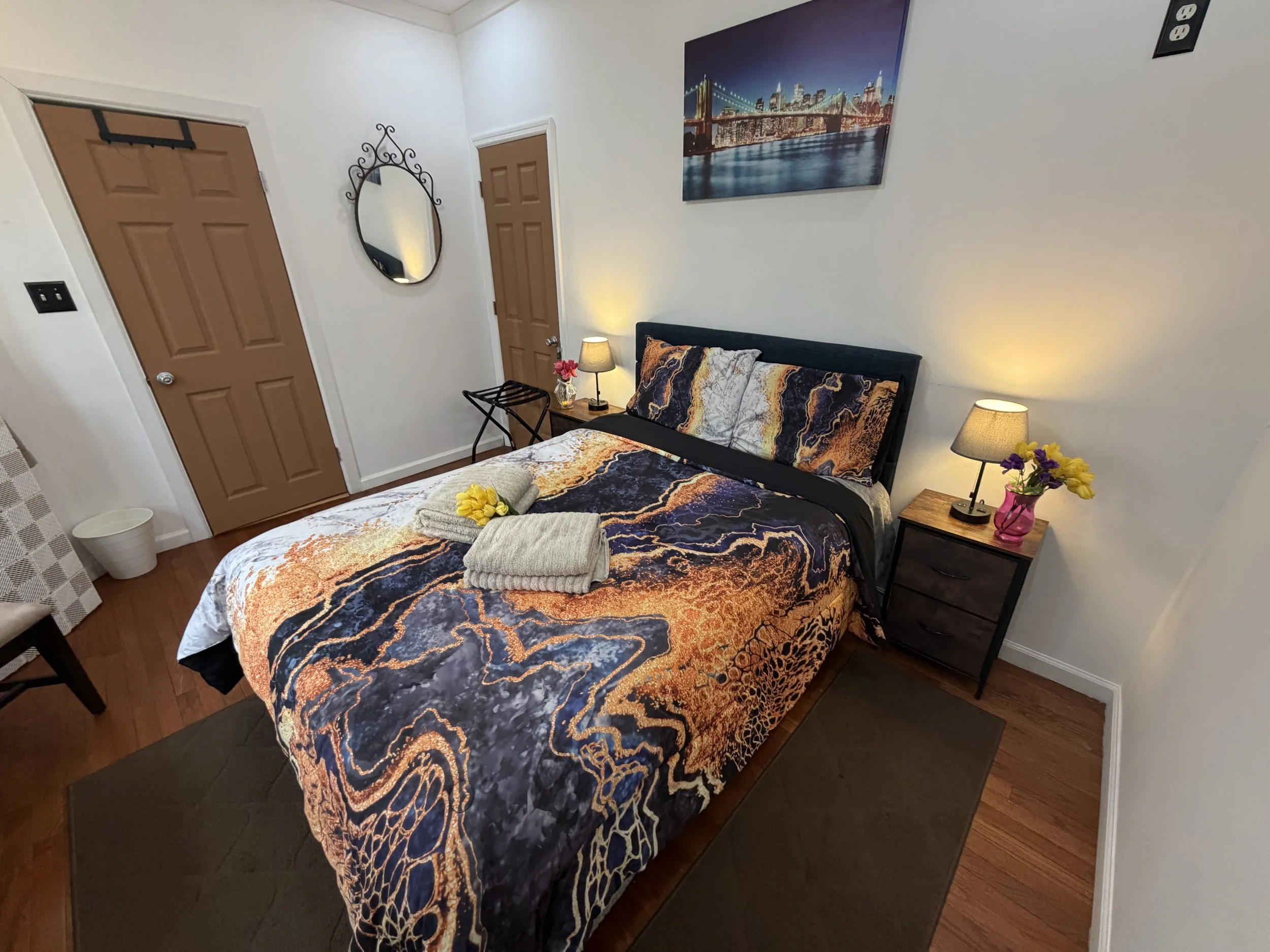 A cozy bedroom with a bed covered in a vibrant, abstract-patterned bedspread in navy, orange, and white. There are folded towels and yellow flowers on the bed. Two bedside tables with lamps and yellow flowers are on either side of the bed. A large cityscape painting hangs on the wall, and a door with a mirror and a wall hook are visible.