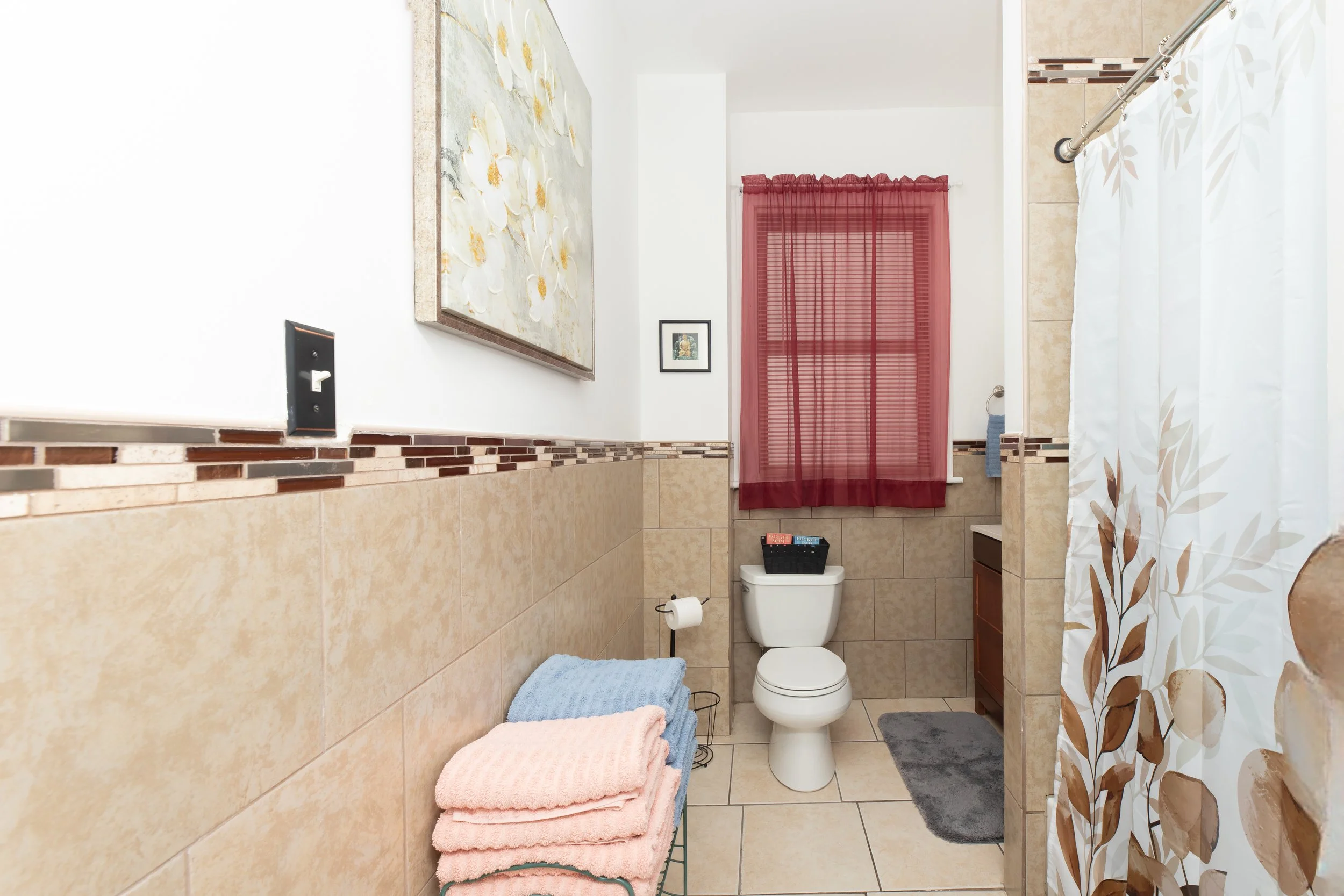 A small bathroom with beige tiles on the lower walls, a toilet, a window with red curtains, a stack of colored towels, a shower curtain with a leaf pattern, and some wall art.