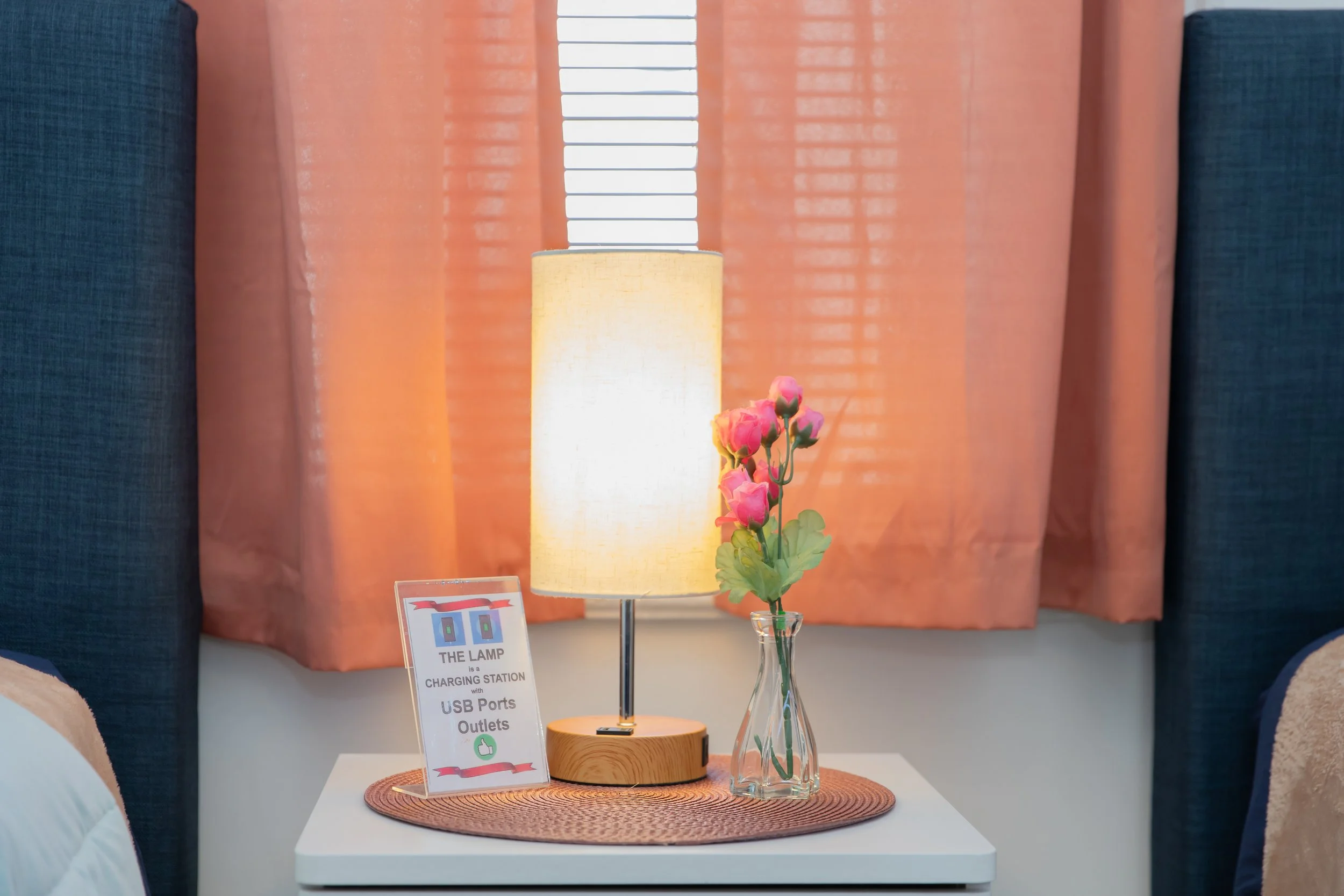 A bedside table with a lamp and a pink flower in a glass vase, in front of closed orange curtains with blinds behind.