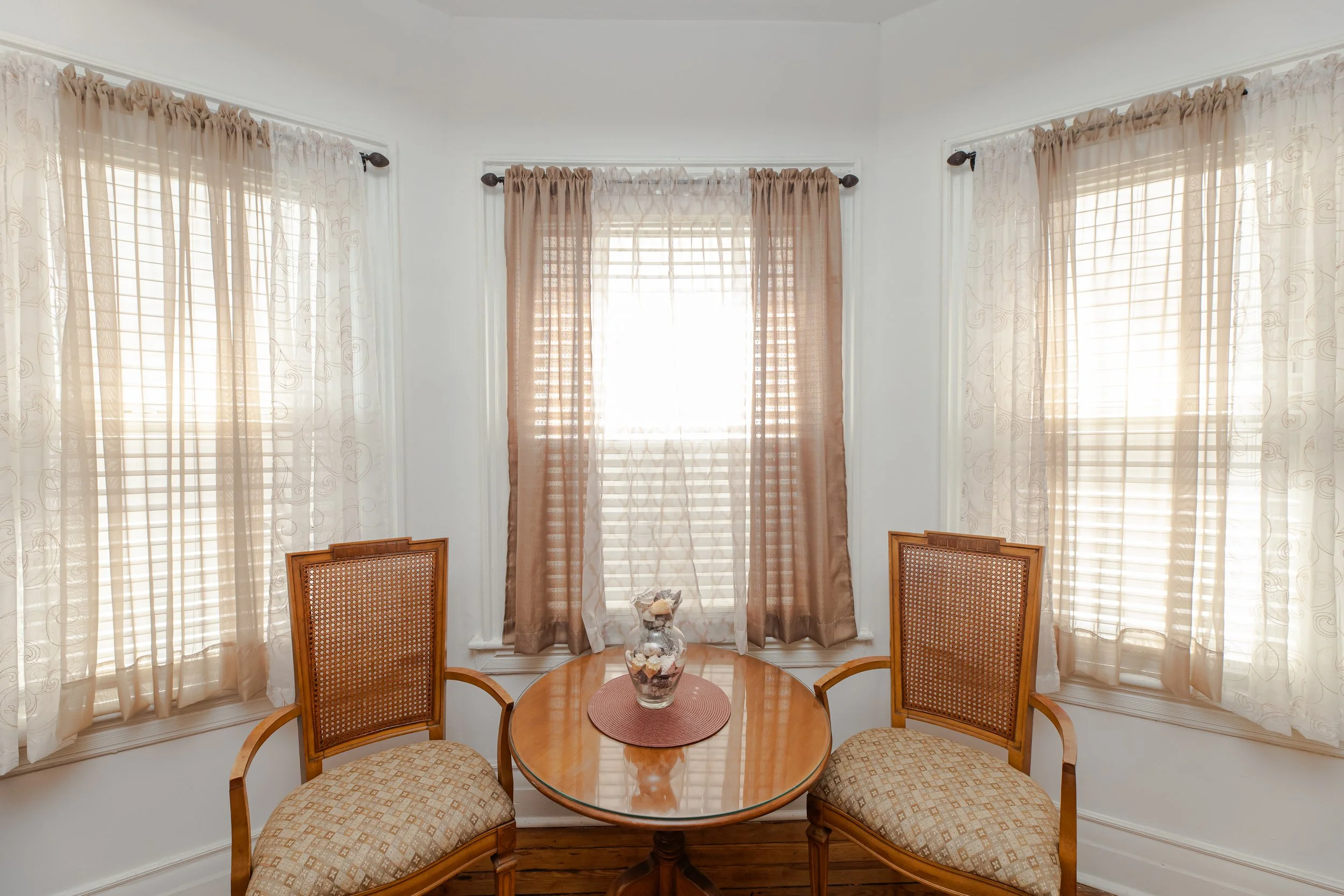 A small round wooden table with a glass vase in the center, flanked by two wooden chairs with patterned fabric cushions, positioned in front of three windows with beige and cream curtains in a bright room.