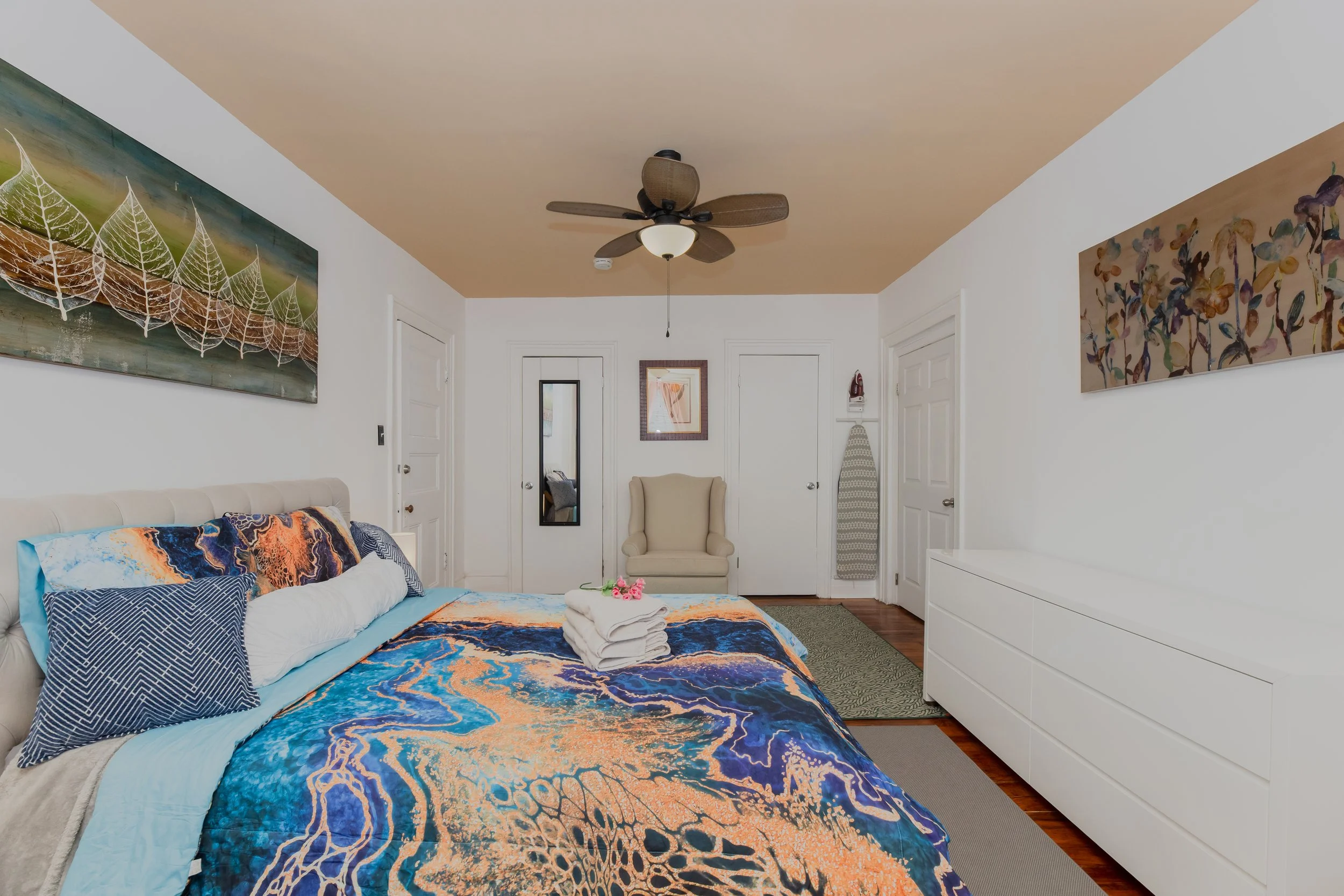 A bedroom with white walls, a bed with colorful abstract-style bedding and pillows, a ceiling fan, a large white dresser, wall art, an armchair, and an ironing board.