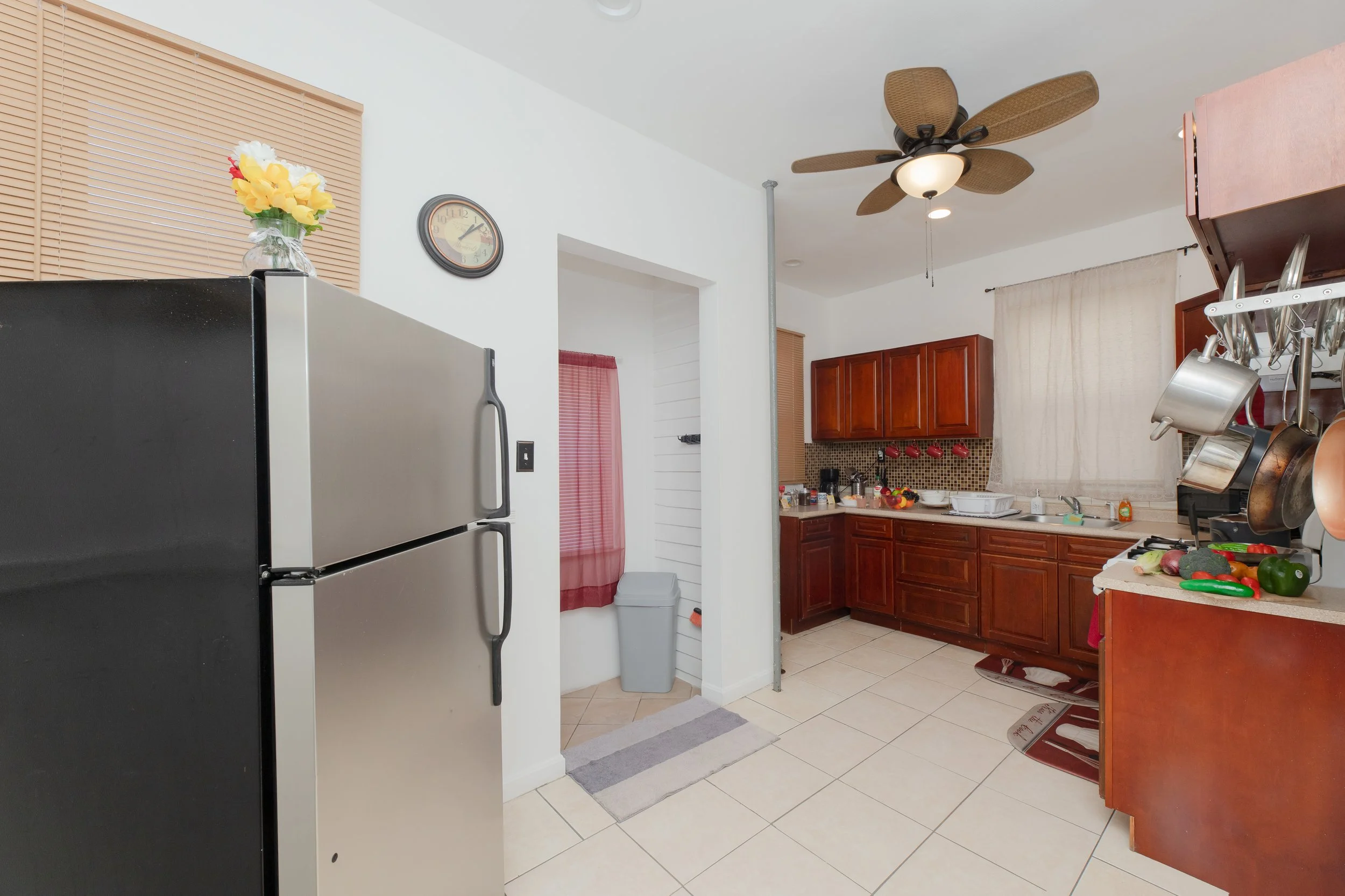Kitchen with wooden cabinets, a silver refrigerator, a ceiling fan with wood blades, kitchenware on a rack, a window with curtains, a tiled floor, and various kitchen items on the counters.