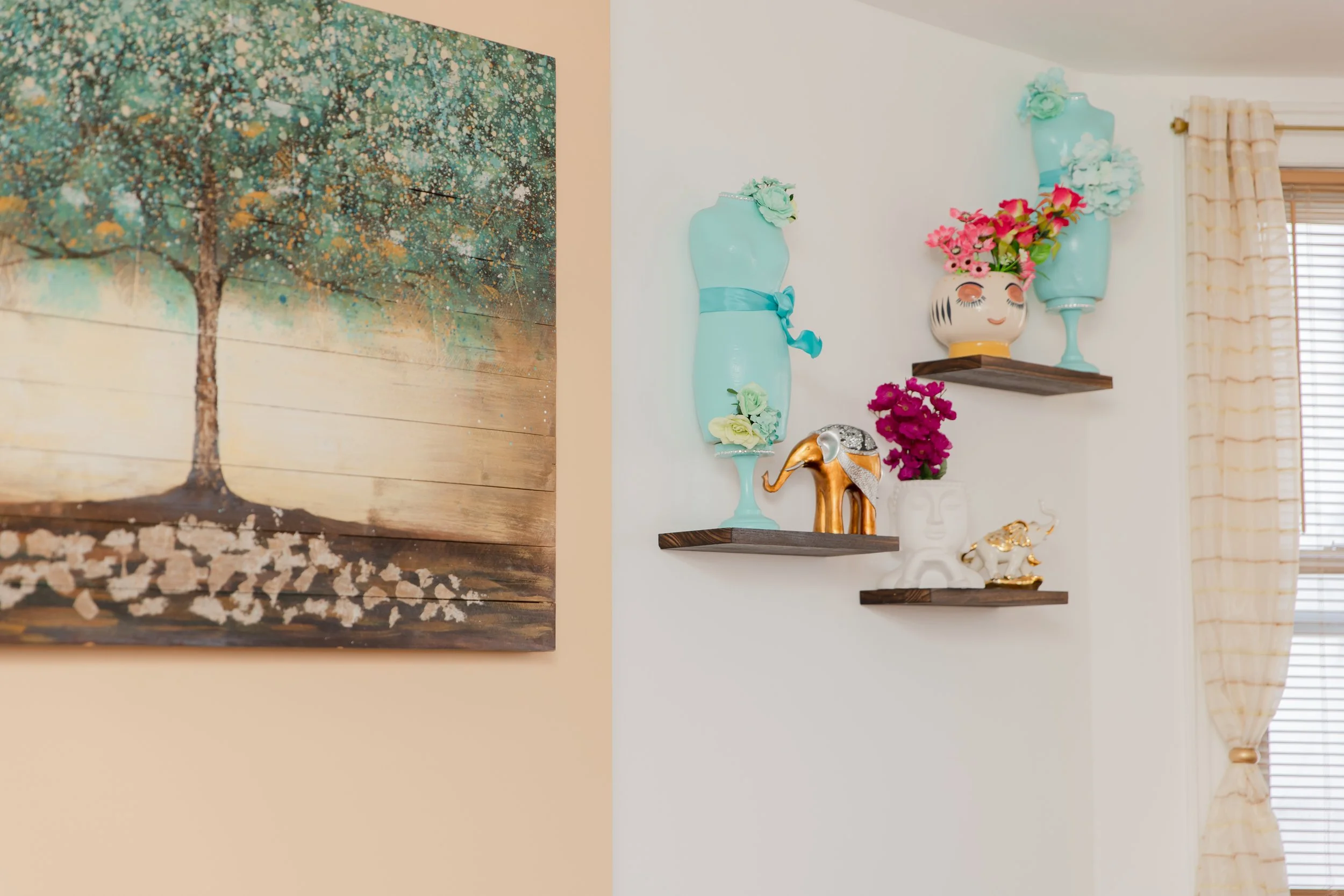 Interior wall decorated with colorful vases, a painted gold elephant figurine, a white face mask sculpture, and pink flowers, with a large abstract tree painting on the adjacent wall and a window with beige curtains on the right.