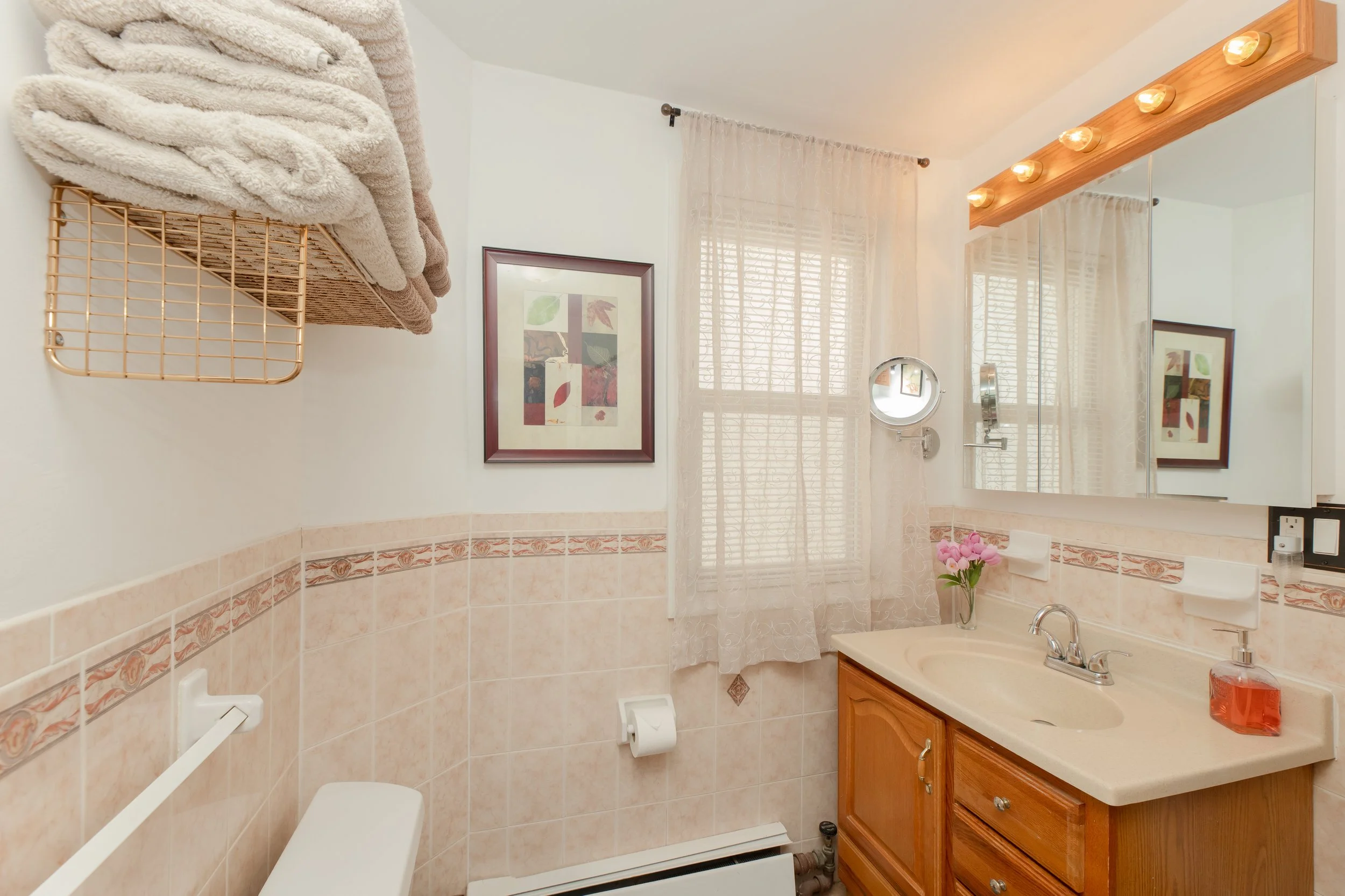 Small bathroom with beige tiles, wooden vanity, mirror, pink flowers, and window with lace curtains.