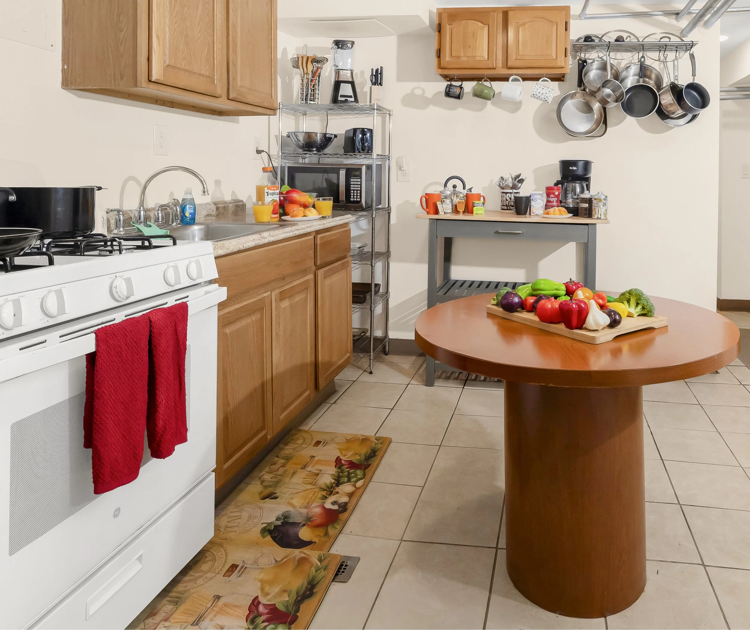 A kitchen with wooden cabinets, a white stove with red towels hanging, a tiled floor, and a round wooden table with fresh vegetables on a cutting board.