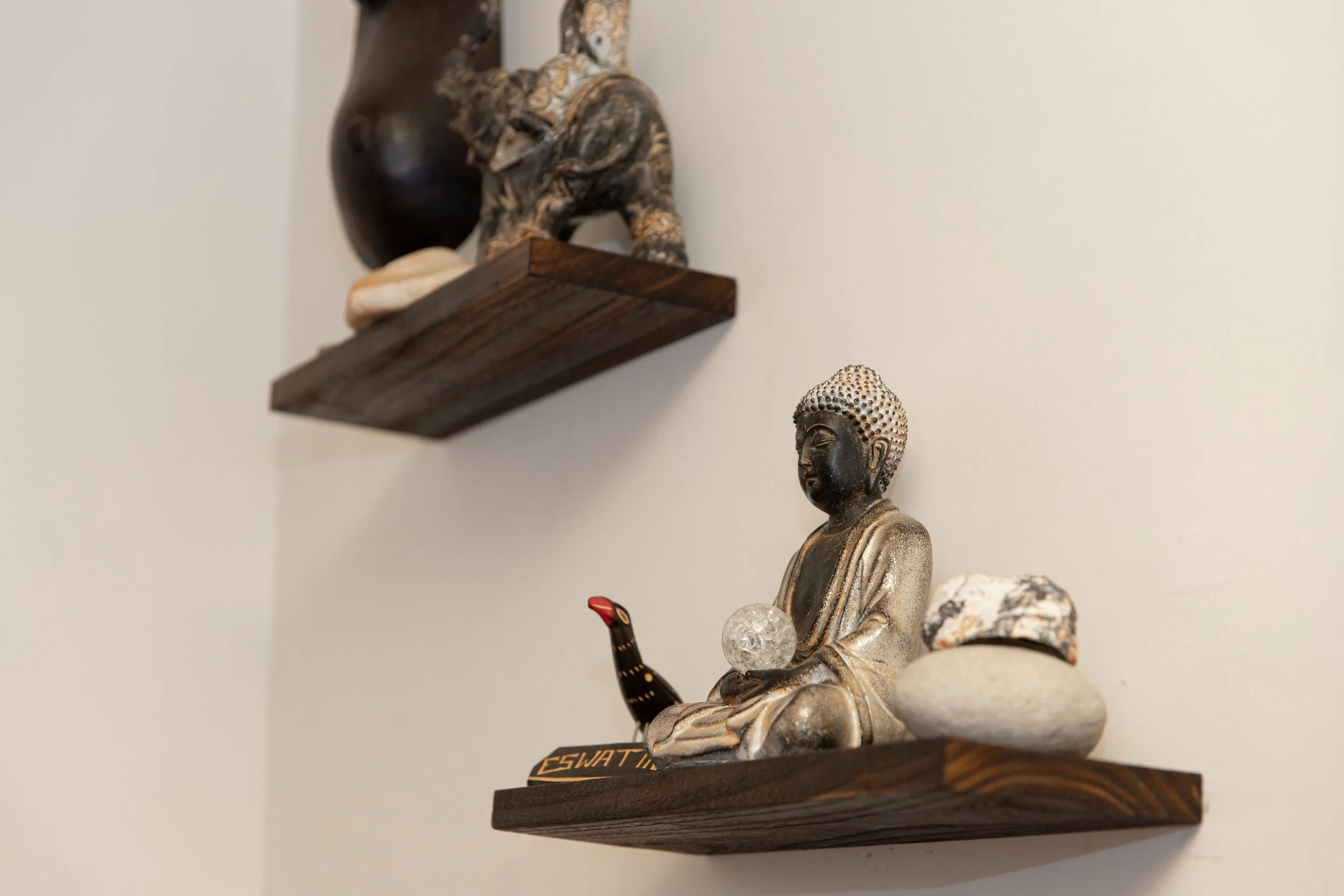 Decorative Buddha statue with a crystal ball and a small painted bird figurine on a wooden shelf, with another wooden shelf and decorative objects above.