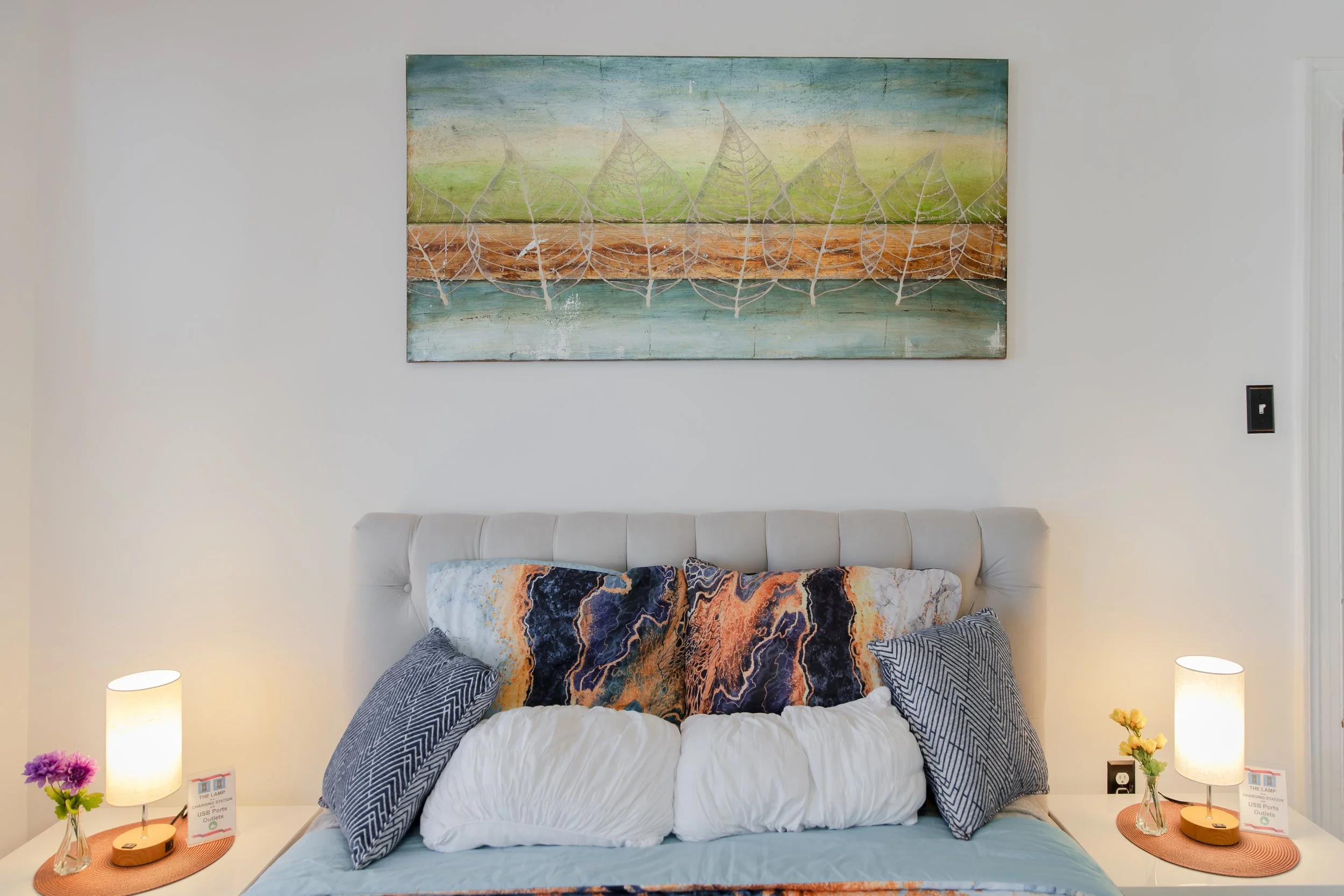 A bedroom with a white padded headboard, decorative pillows, and two side tables with lamps and flowers, and a large abstract painting above the bed.