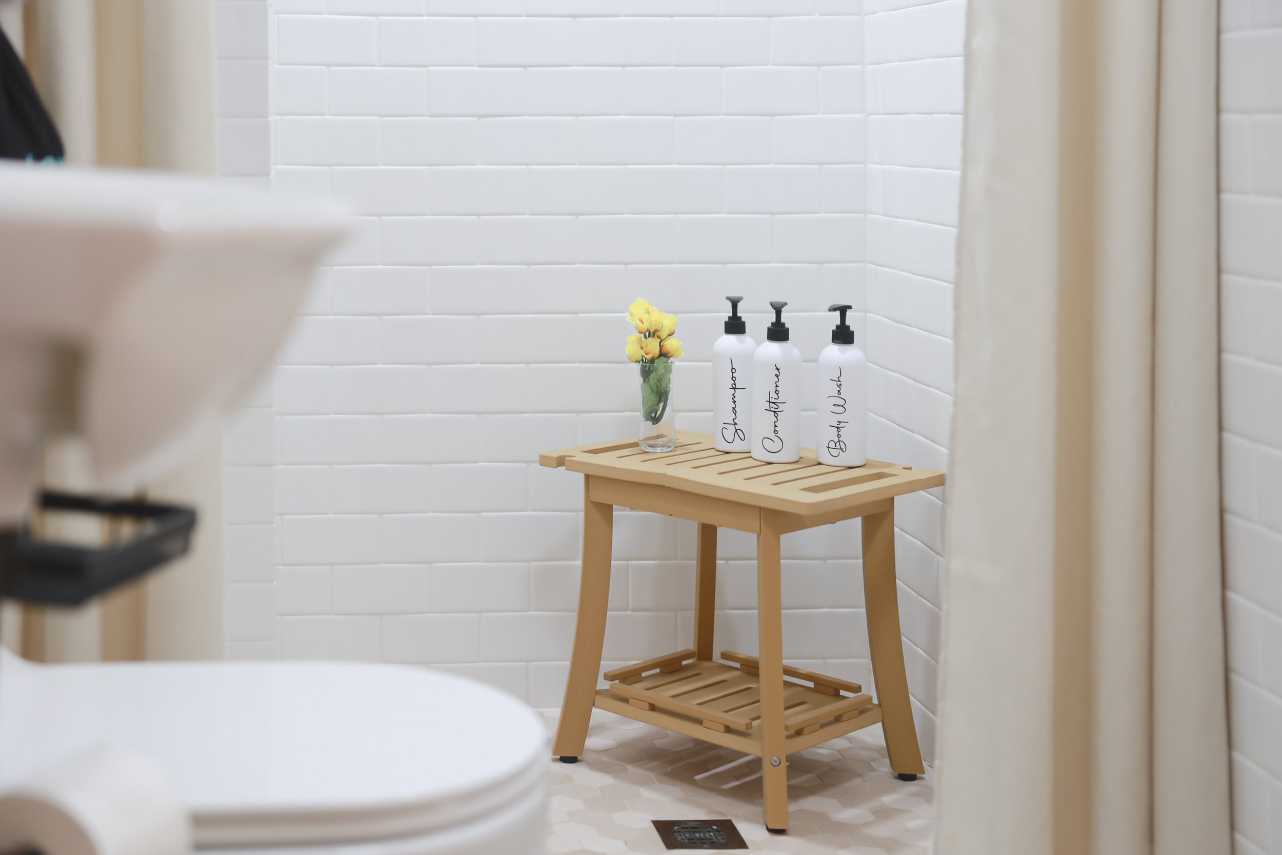A wooden table with three white bottles labeled shampoo, conditioner, and body wash, and a small flower vase, in a bathroom with white tiled walls.